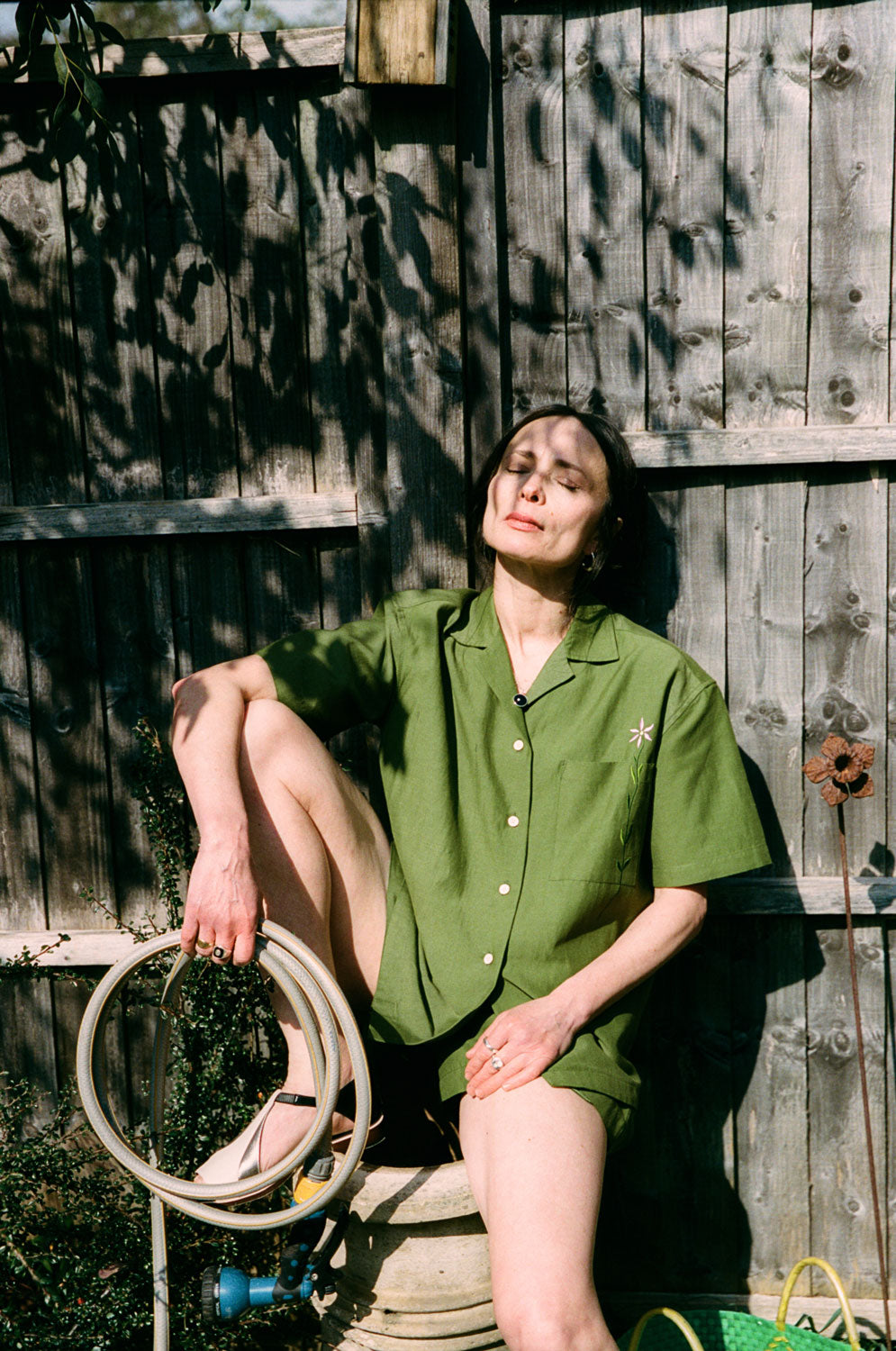 A woman is perched on a garden pot, holding a garden hose, with a wooden fence behind her, with leafy shadows on the fence. She is wearing the Frances Unisex Cuban Shirt in olive green, with short sleeves and the signature embroidered flower in lilac on the check pocket. Worn with the Bessie A-line Shorts in olive green as a matching twin set. She is holding a curled up garden hose, with one leg up on the garden chimney pot.