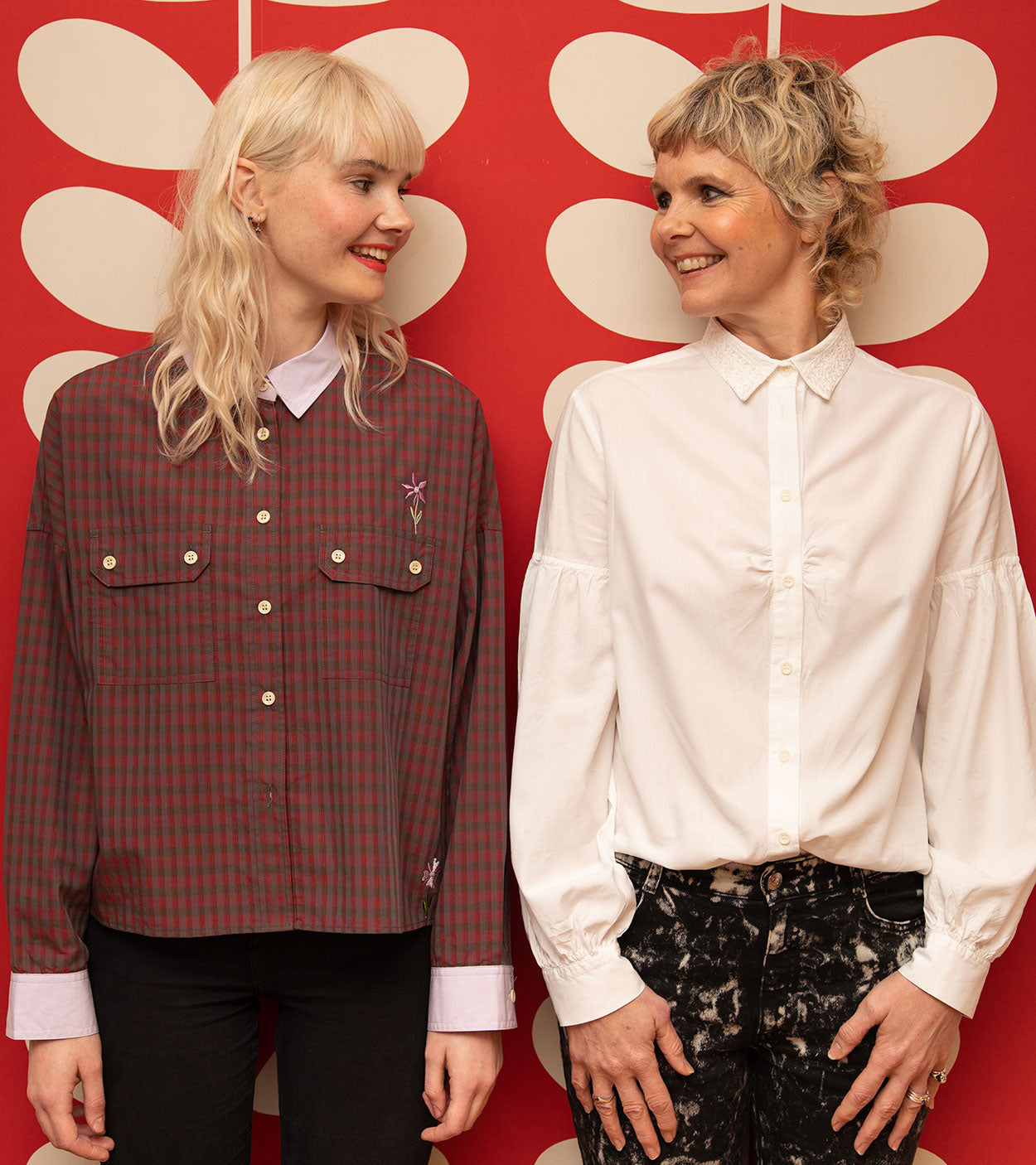 A mum and her daughter stand side by side, smiling at each other. The mum is wearing Saywood's white Edi Volume Sleeve Shirt with lace collar, and the daughter is wearing Saywood's Jules Utility Shirt in micro red check with lilac collar. A red and white wallpaper is in the background.