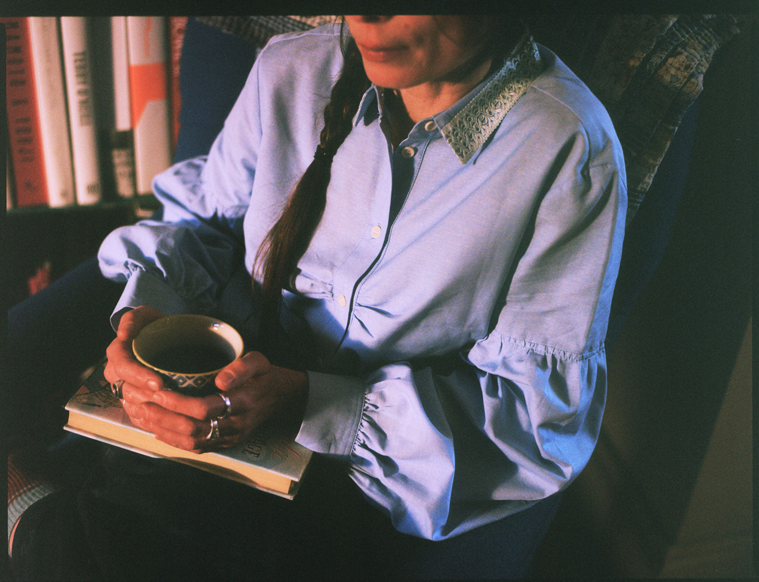 Close up of a woman sitting in a blue antique armchair, holding a cup of tea with a book in her lap. She is wearing Saywood's Edi Volume Sleeve Shirt with lace trim collar in pale blue recycled cotton. The soft gathers at the bust can be seen on the shirt. A bookcase can be seen behind her.