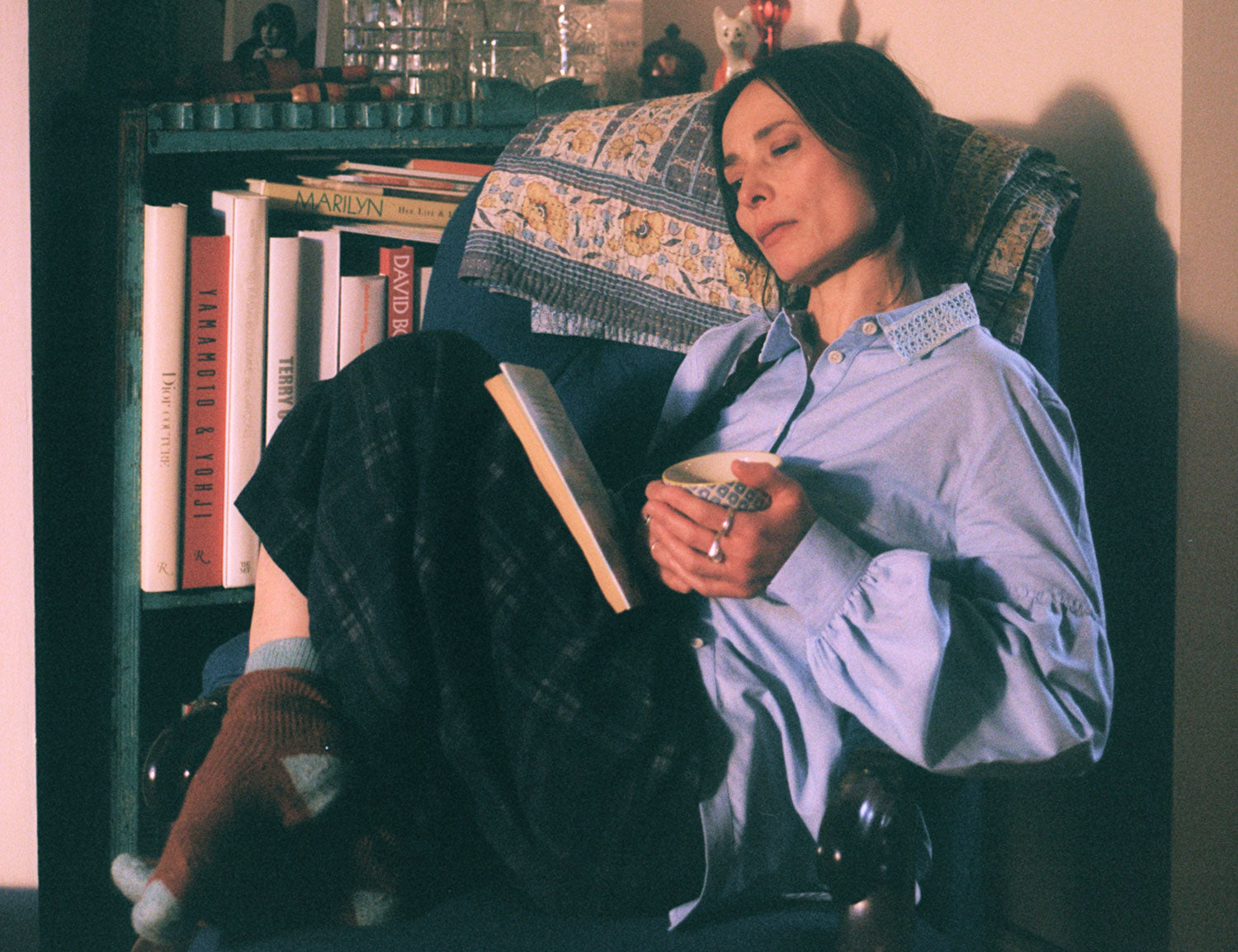 Close up of a woman sitting in a blue antique armchair, with her knees up, holding a cup of tea with a book in her lap. She is wearing Saywood's Edi Volume Sleeve Shirt with lace trim collar in pale blue recycled cotton, with the Amelia Wide Leg Trousers in navy check, and cosy brown and blue socks. A bookcase is behind her, with a lamp and ornaments on.