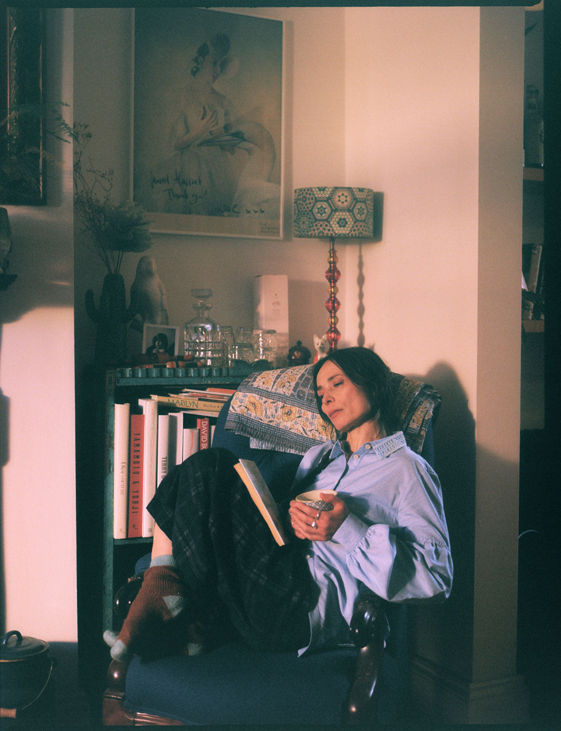 A woman sits in a blue antique armchair, with her knees up, holding a cup of tea with a book in her lap. She is wearing Saywood's Edi Volume Sleeve Shirt with lace trim collar in pale blue recycled cotton, with the Amelia Wide Leg Trousers in navy check, and cosy brown and blue socks. A bookcase is behind her, with a lamp and ornaments on, with a picture by artist Linder Sterling on the wall.