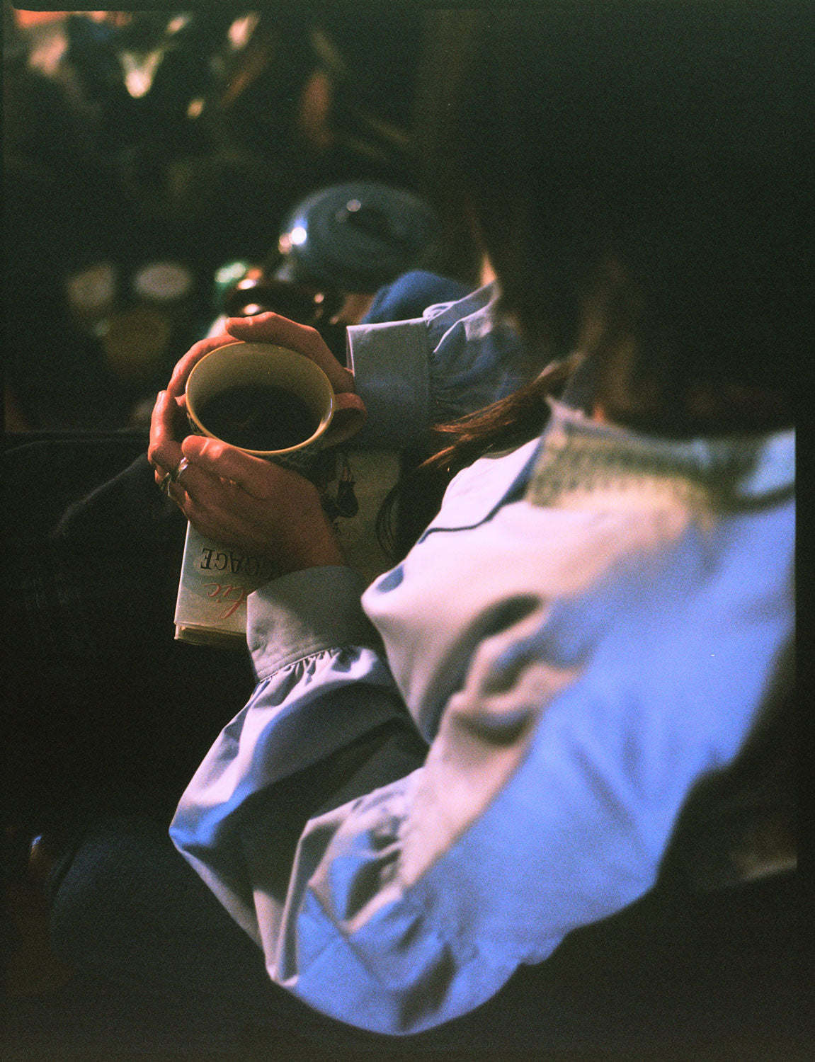 A woman sits in a blue antique armchair, holding a cup of tea with a book in her lap. She is wearing Saywood's Edi Volume Sleeve Shirt with lace trim collar in pale blue recycled cotton. The image is shot over her shoulder.