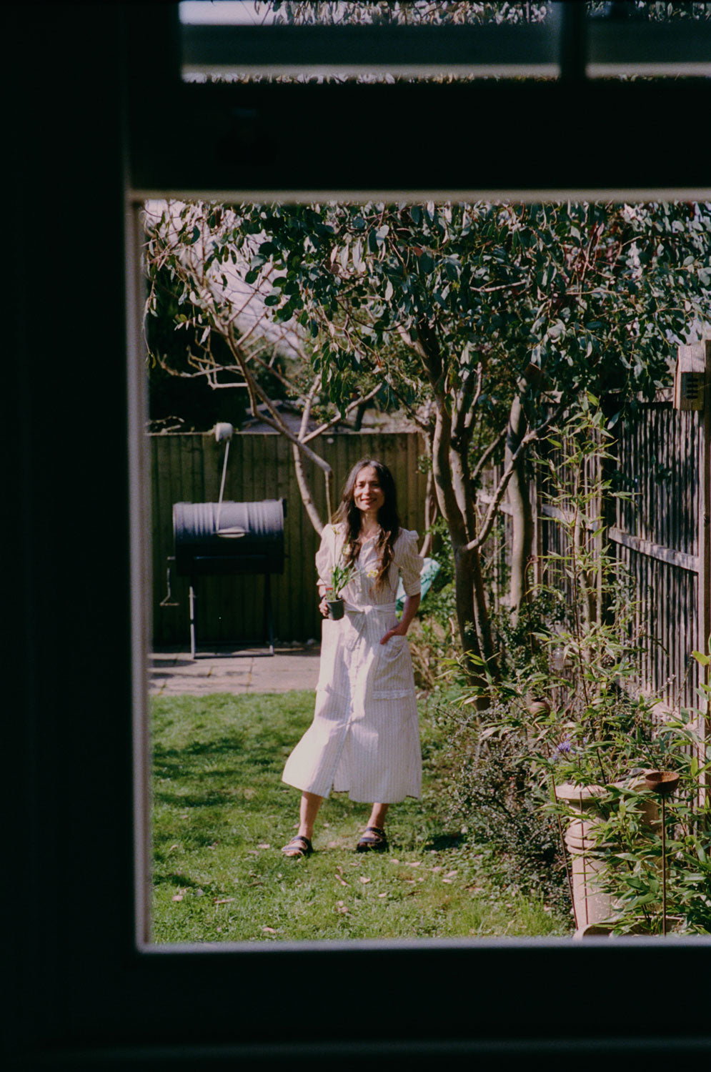A woman stands in a garden, by a eucalyptus tree, holding a daffodil plant. She is wearing Saywood's white cotton pinstripe Rosa Puff Sleeve Shirtdress with the tie belt tied round her waist, with the patch pockets with the ruffle trim just visible. She wears her hair down, with black sandals on her feet, and one hand in her pocket. The image is shot through an open window.