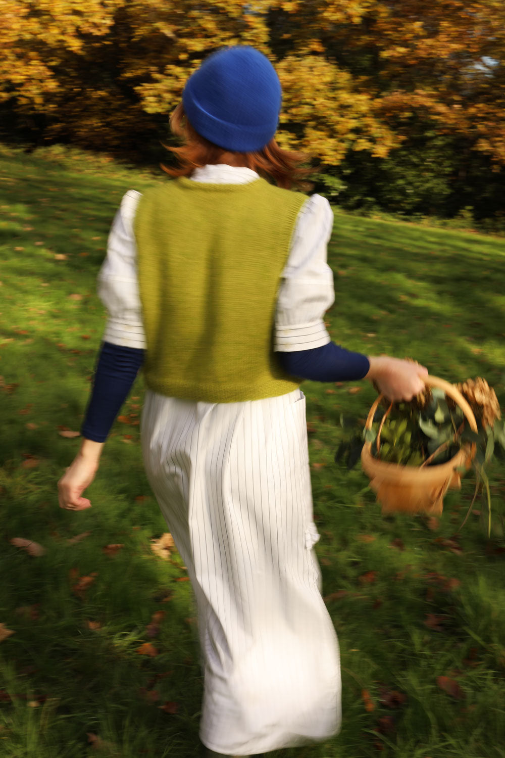 Woman is walking with her back to the camera, surrounded by trees, holding a wooden garden basket with flowers in. She is wearing Saywood's white pinstripe shirtdress, the Rosa Puff Sleeve Shirtdress, with a navy long sleeve top underneath and a green sweater vest layered over the top, with a blue beanie hat. The image is slightly blurred as she moves.