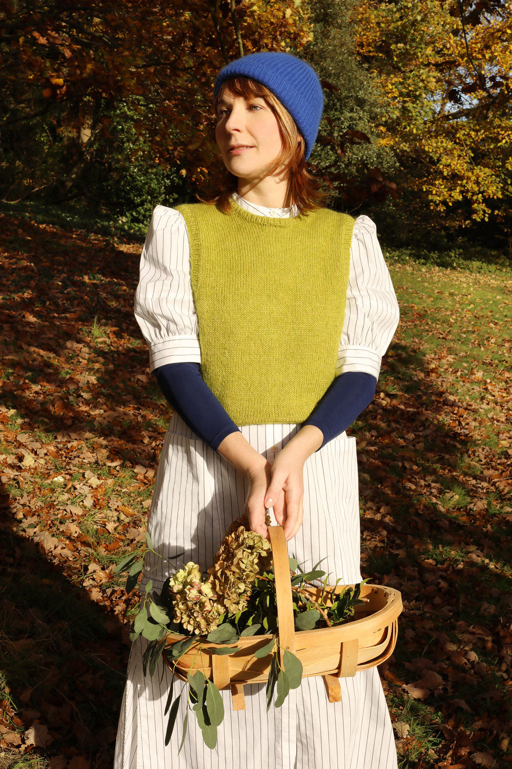 Woman stands surrounded by trees, holding a wooden garden basket with flowers in. She is wearing Saywood's white pinstripe shirtdress, the Rosa Puff Sleeve Shirtdress, with a navy long sleeve top underneath and a green sweater vest layered over the top, with a blue beanie hat.