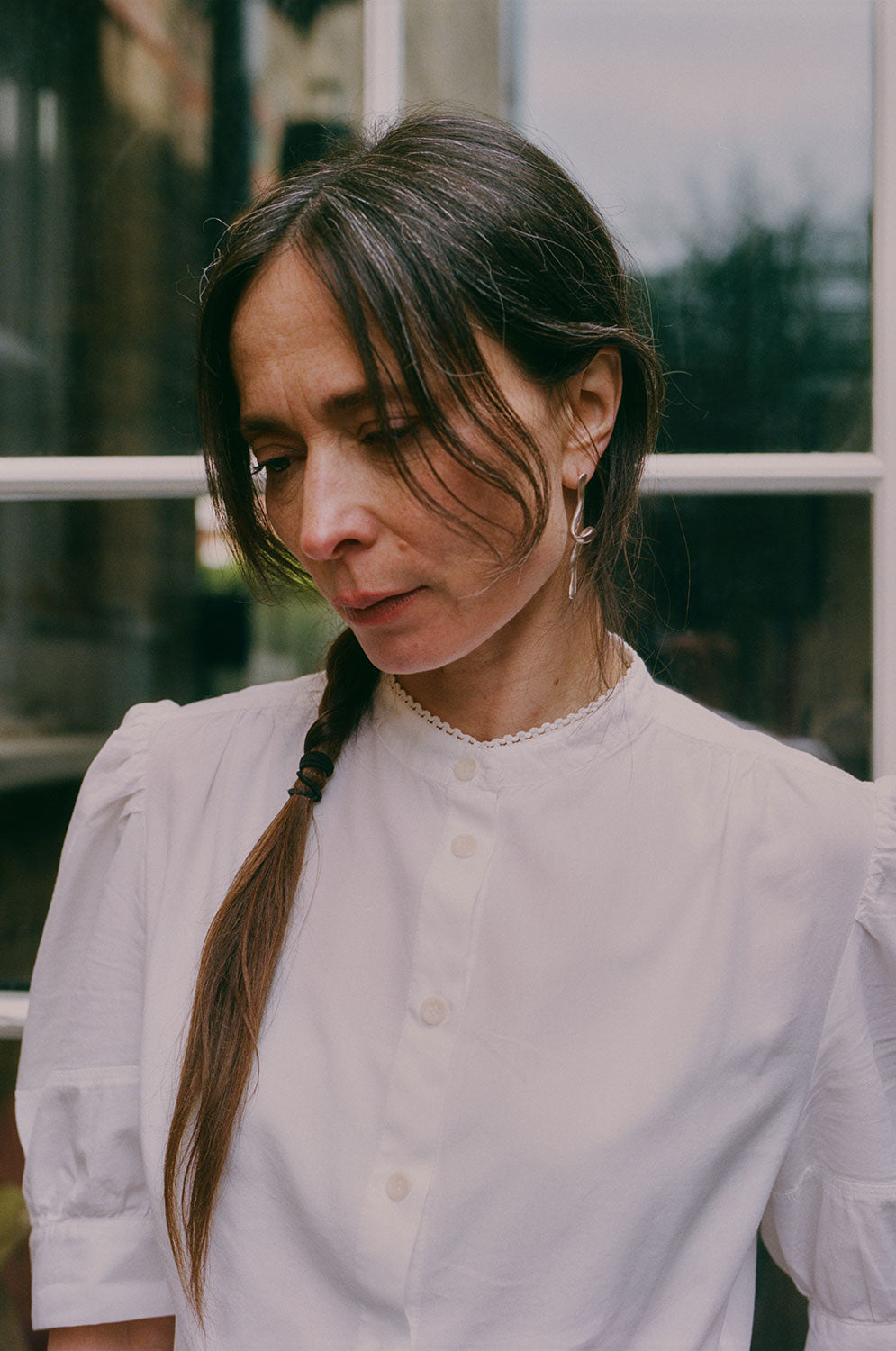 Close up of a woman with dark hair, plaited, looking to one side. She is wearing Saywood's Joni Puff Sleeve Blouse, with a silver twisted earring in her ear. The buttons down the blouse can be seen, as well as the grandad collar with lace trim on the edging, and puff sleeve detailing.