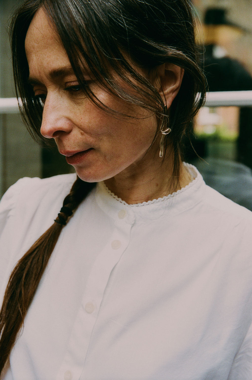 Close up of a woman with dark hair looking to one side. She is wearing Saywood's Joni Puff Sleeve Blouse, with a silver twisted earring in her ear. The buttons down the blouse can be seen, as well as the grandad collar with lace trim on the edging.