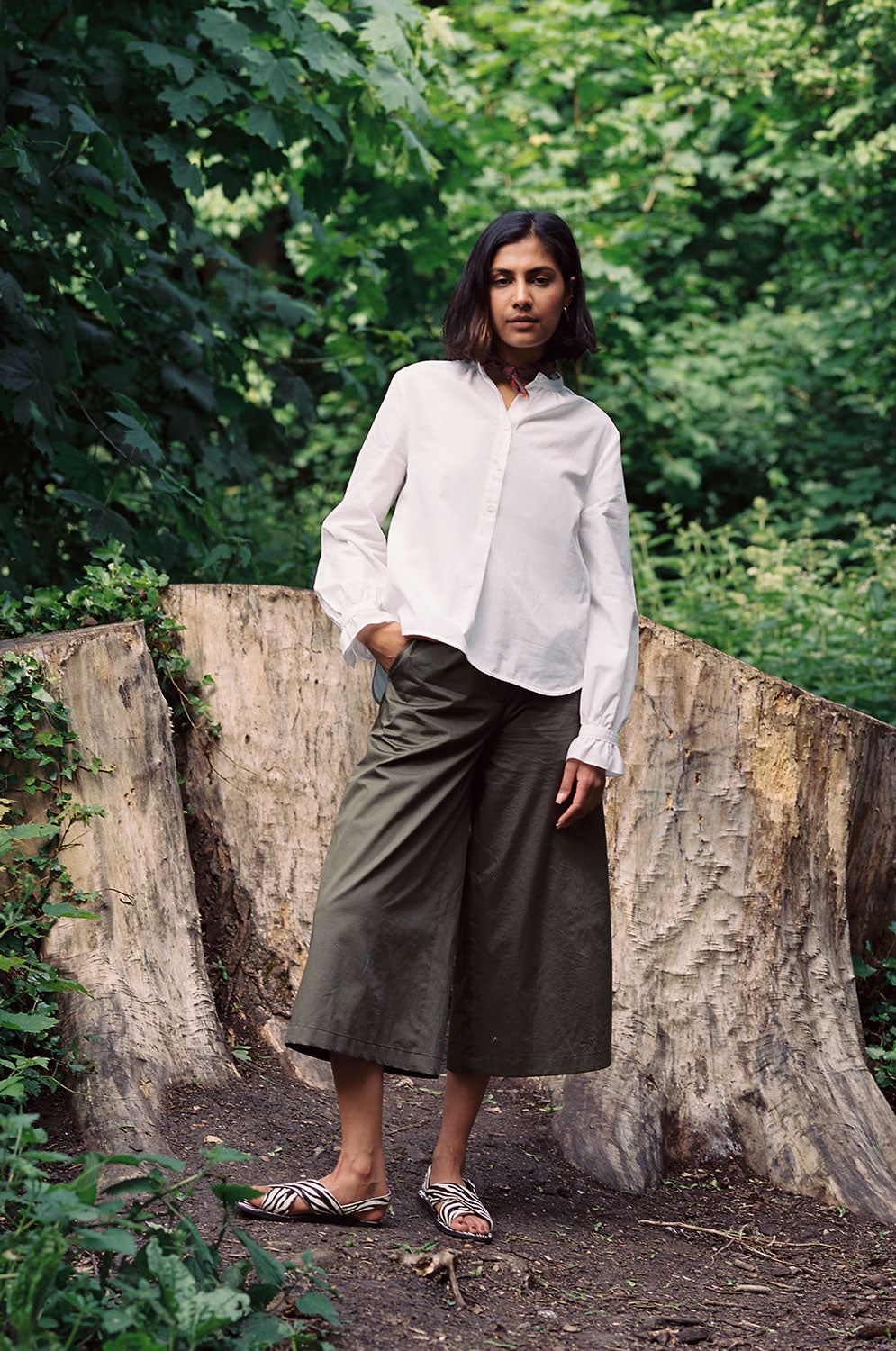 Model stands, legs in wide stance, in front of tree stumps and greenery. She wears Saywood's white Marie A-line Blouse with a red check neckerchief tied round her neck. Worn with Saywood's Amelia wide leg trouser in khaki and black and white zebra print sandals.