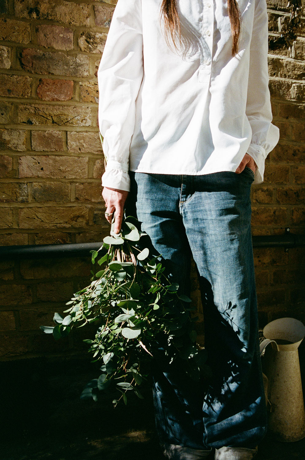 A woman standing in front of a brick wall, holding a bunch of eucalyptuses. The shot is from the shoulders down. She is wearing rings on her fingers, and jeans, worn with Saywood's white recycled cotton Marie A-line Gathered Neck blouse. the sleeve detail of the gathered cuff and frill with lace trim can be seen. Dappled light and leafy shadows are seen on the blouse. A vintage white jug can be seen on the floor.