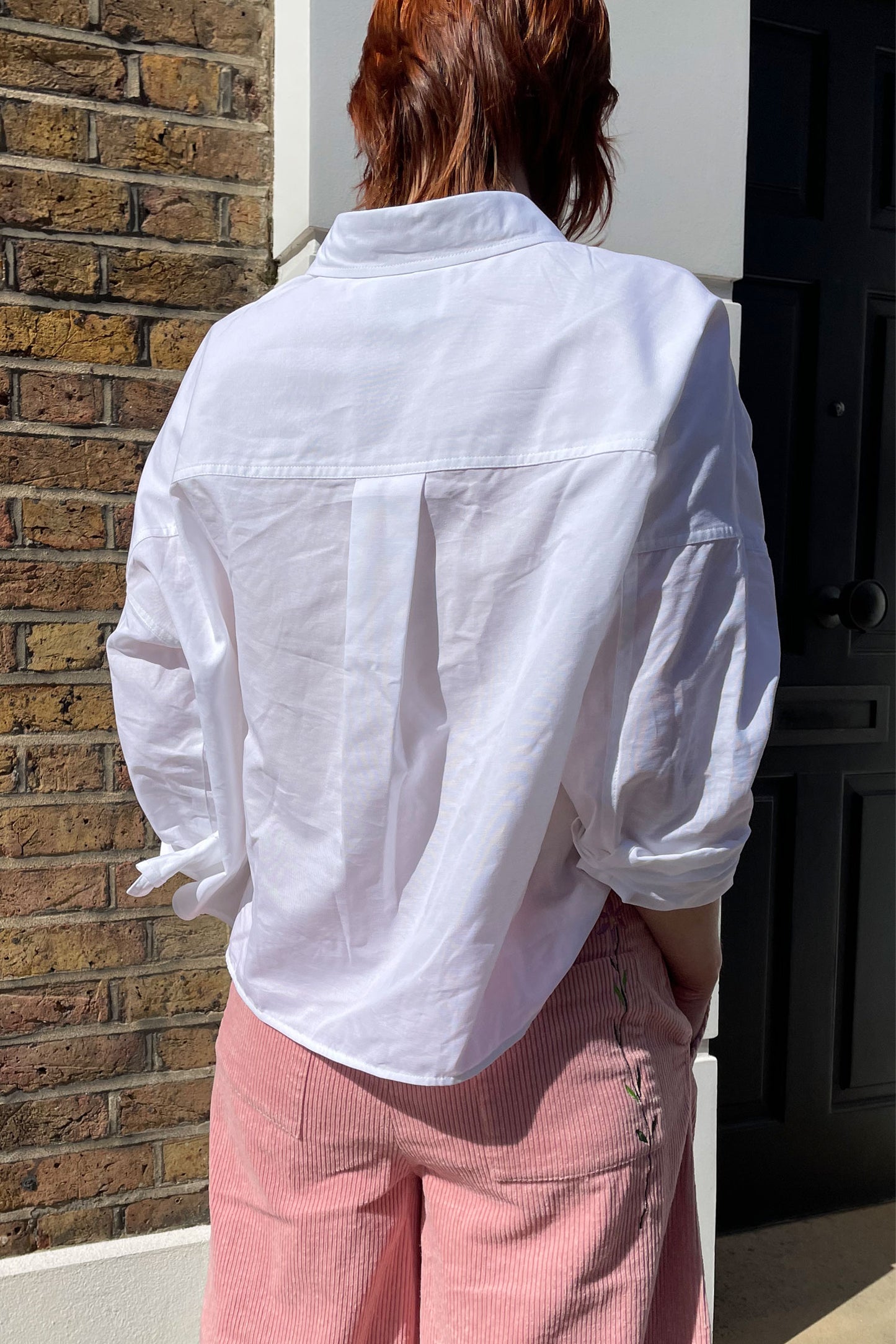 Woman stands with her back to the camera. She wears Saywood's white recycled cotton women's boxy Lela Shirt. The back box pleat can be seen. She wears a pair of pink corduroy Amelia Trousers with the Saywood signature flower embroidery on the back pocket. Made in London