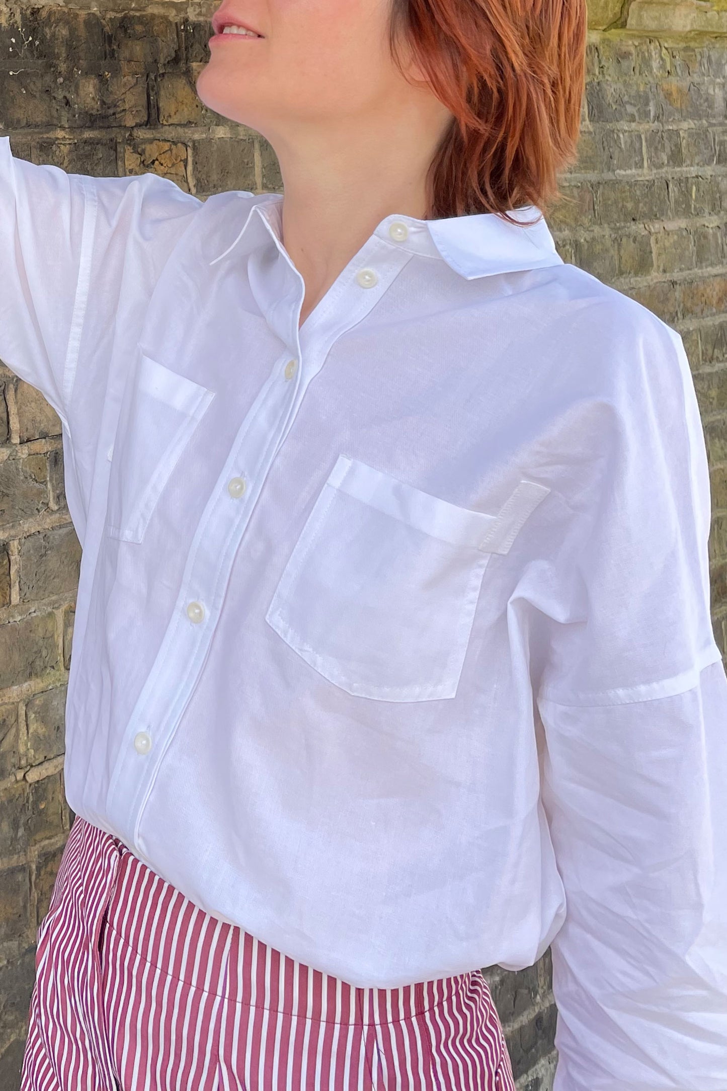 Close up of the woman wearing the Saywood boxy fit Lela shirt in white recycled cotton, made in London. The patch pockets and herringbone cotton tape detail can be seen on the pocket corners. Wirn with a pair of red and white stripe shirts.