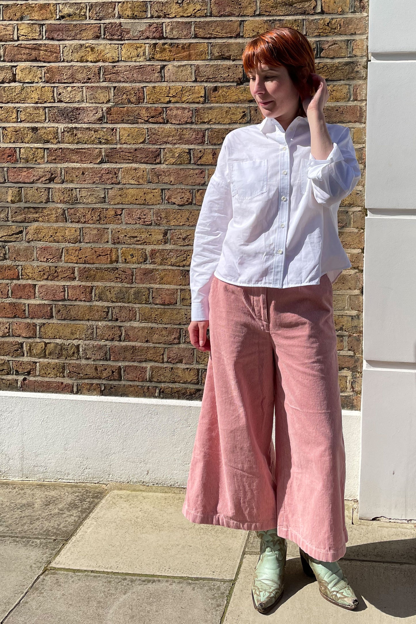 Woman stands in front of a brick wall, wearing Saywood's Lela Boxy Shirt in white recycled cotton. She has her sleeves rolled up with one hand touching her red hair. She wears the pink corduroy Amelia Trouser with pastel green cowboy boots. The shirt is untucked.