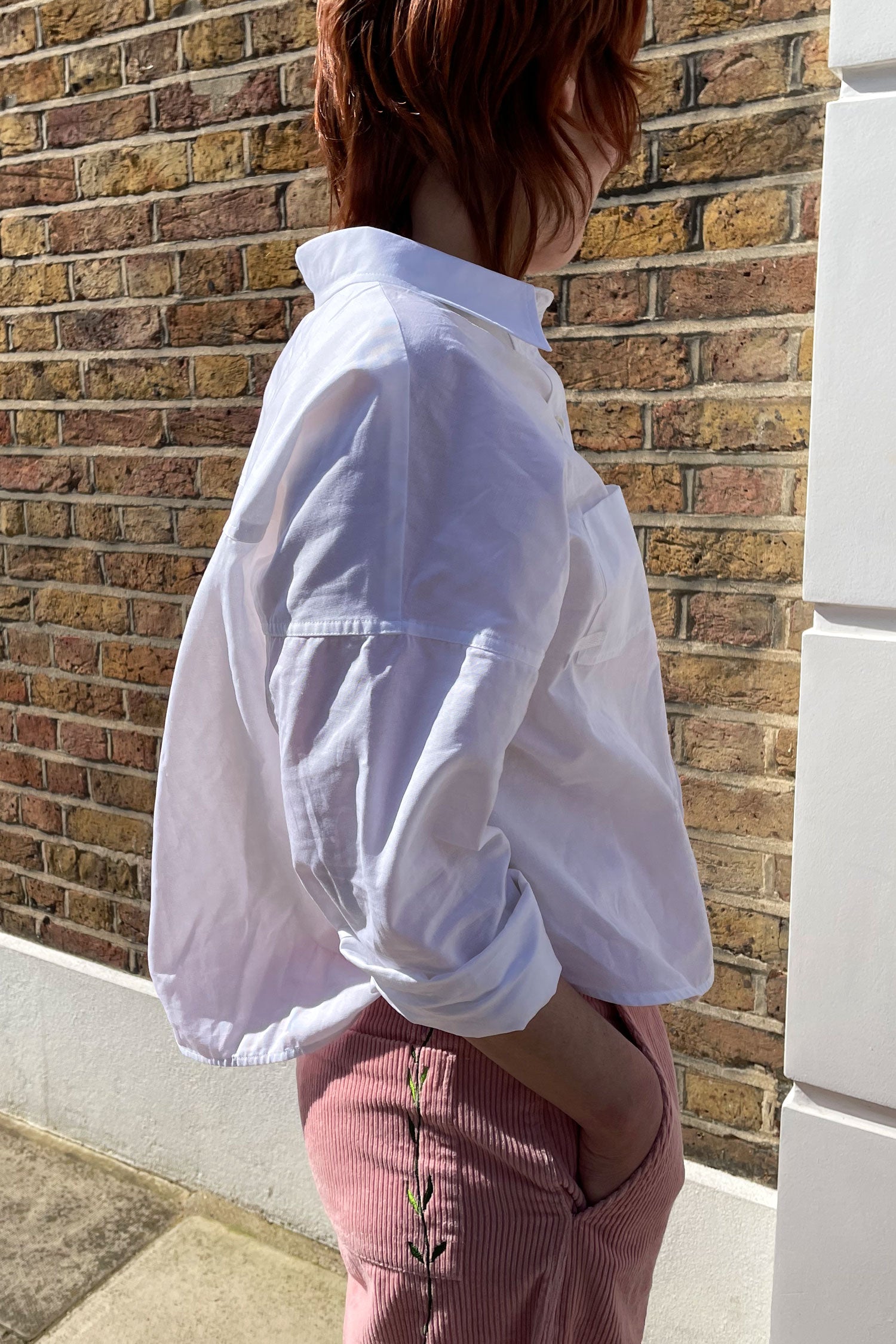 Woman stands side on in front of a brick wall. She wears Saywood's white recycled cotton women's boxy Lela Shirt. The back box pleat can be seen. She wears a pair of pink corduroy Amelia Trousers with the Saywood signature flower embroidery on the back pocket. Made in London