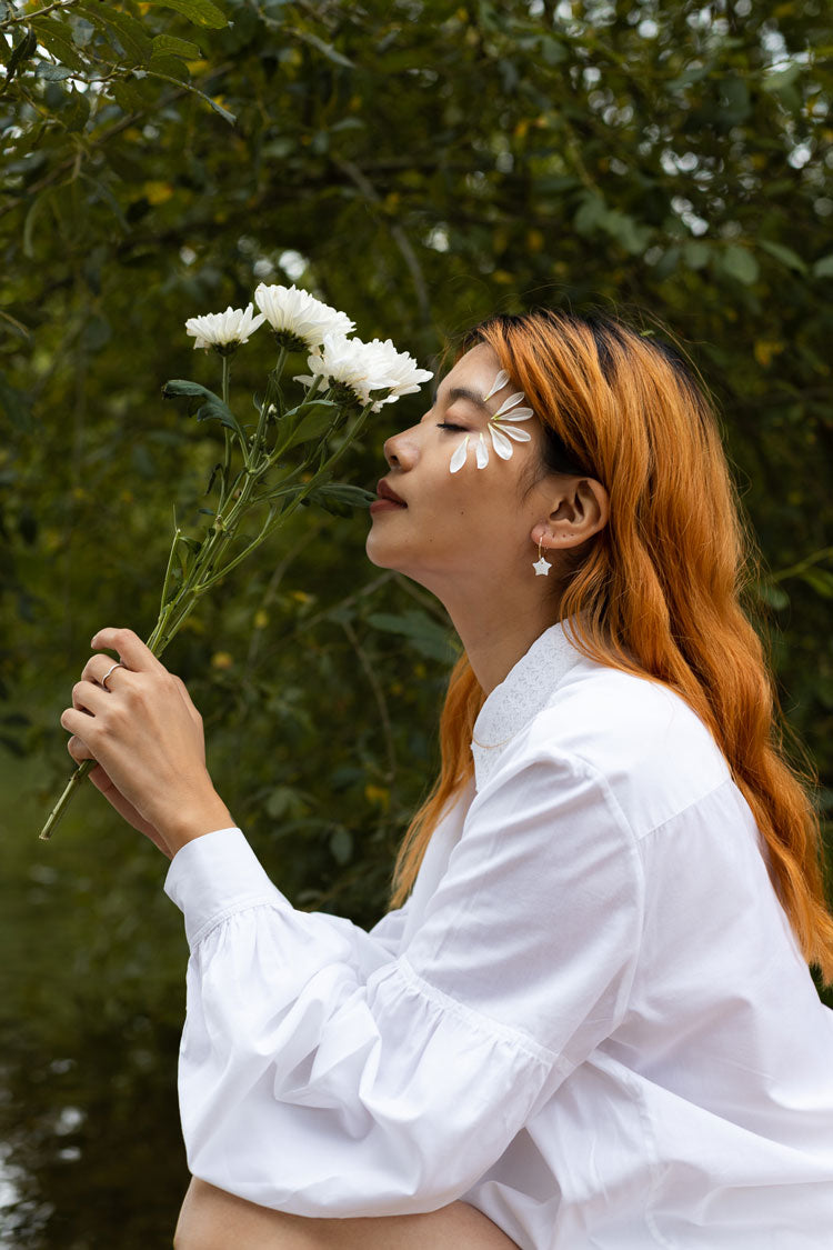 Women's white shirt with lace collar and volume sleeves. Model sits with a flower to her face wearing Saywood's Edi Volume Sleeve Shirt in white Supima cotton and bamboo