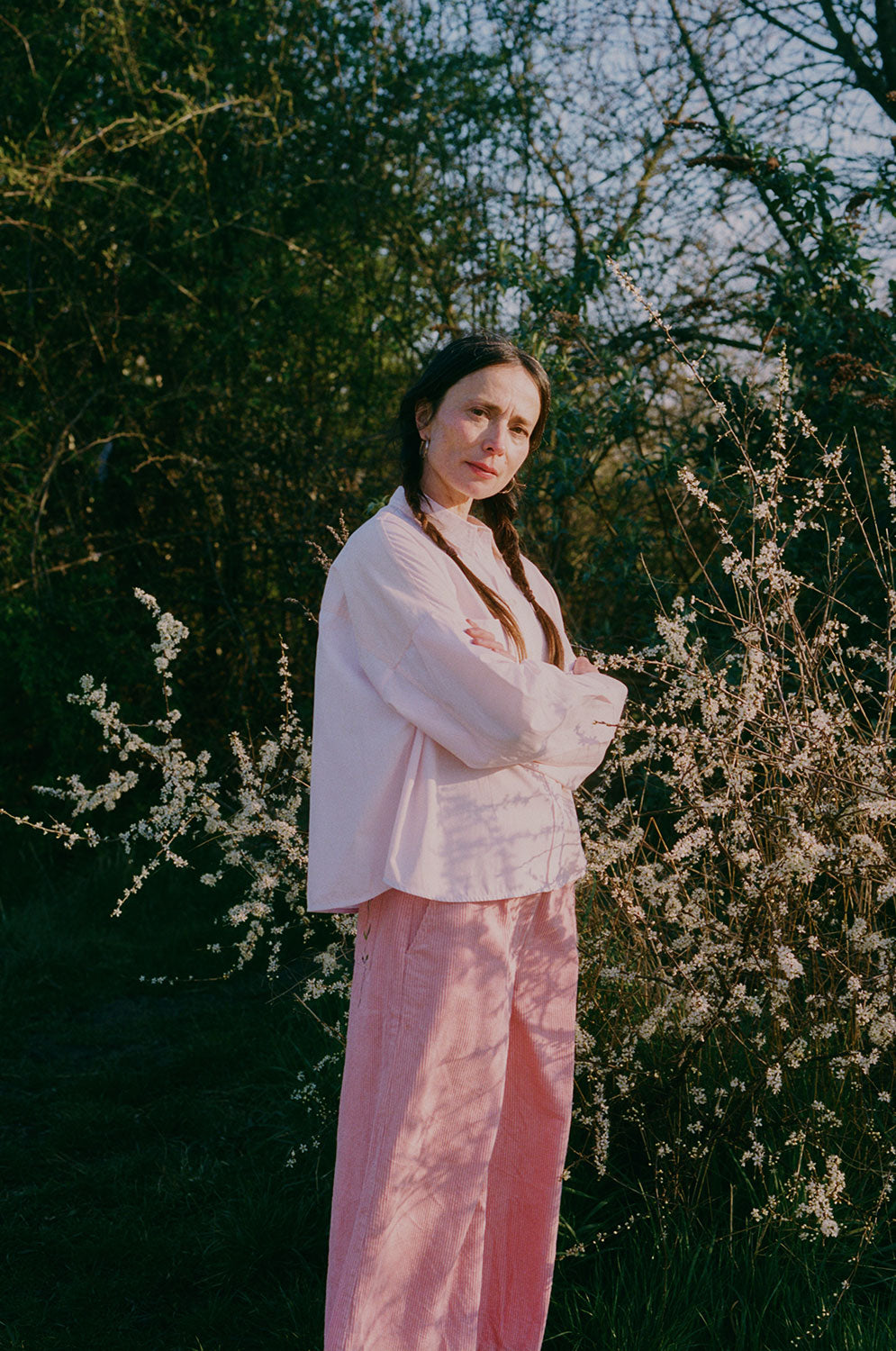 A woman standing amongst the Spring brambles, image shot on film. She wears Saywood's Amelia Wide Leg Trousers in pink corduroy, paired with the Lela Boxy Colourblock Shirt in pastel pink and orange, all made in London. Her hair is in pigtail plaits and her arms are folded across her chest.