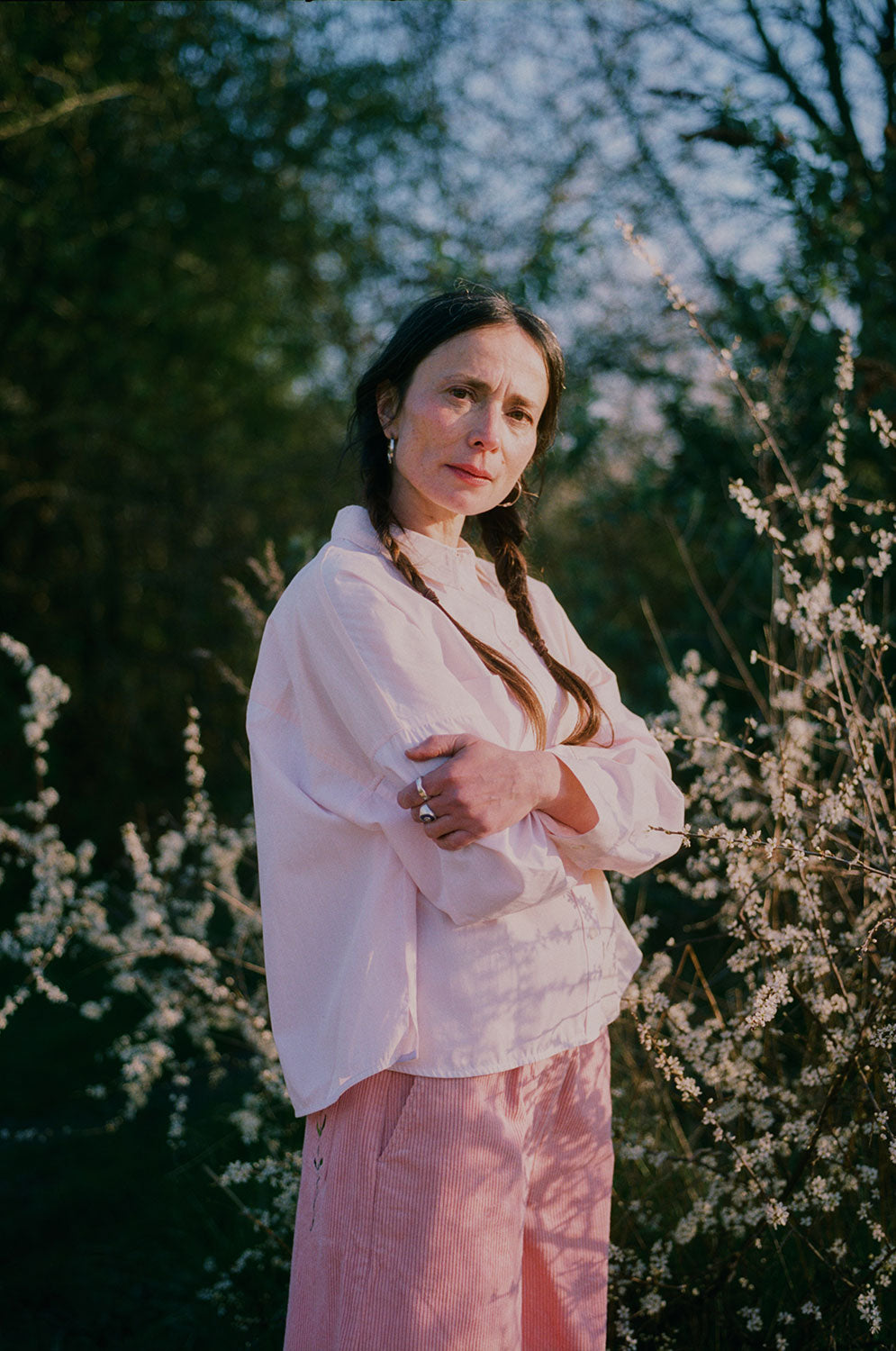 Close up of a woman standing amongst the Spring brambles, image shot on film. She wears Saywood's Amelia Wide Leg Trousers in pink corduroy, paired with the Lela Boxy Colourblock Shirt in pastel pink and orange, all made in London. Her hair is in pigtail plaits and her arms are folded across her chest.