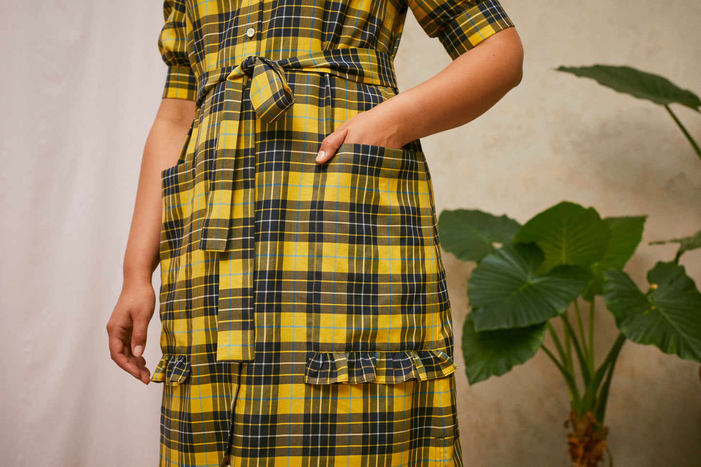 Close up of Saywood's Rosa yellow check shirtdress, with the belt tied at the waist. Model has one hand in the patch pocket, wthe other hangs to her side. Ruffles at the base of the pockets can be seen. A plant and drop of pink fabric can be seen in the background.