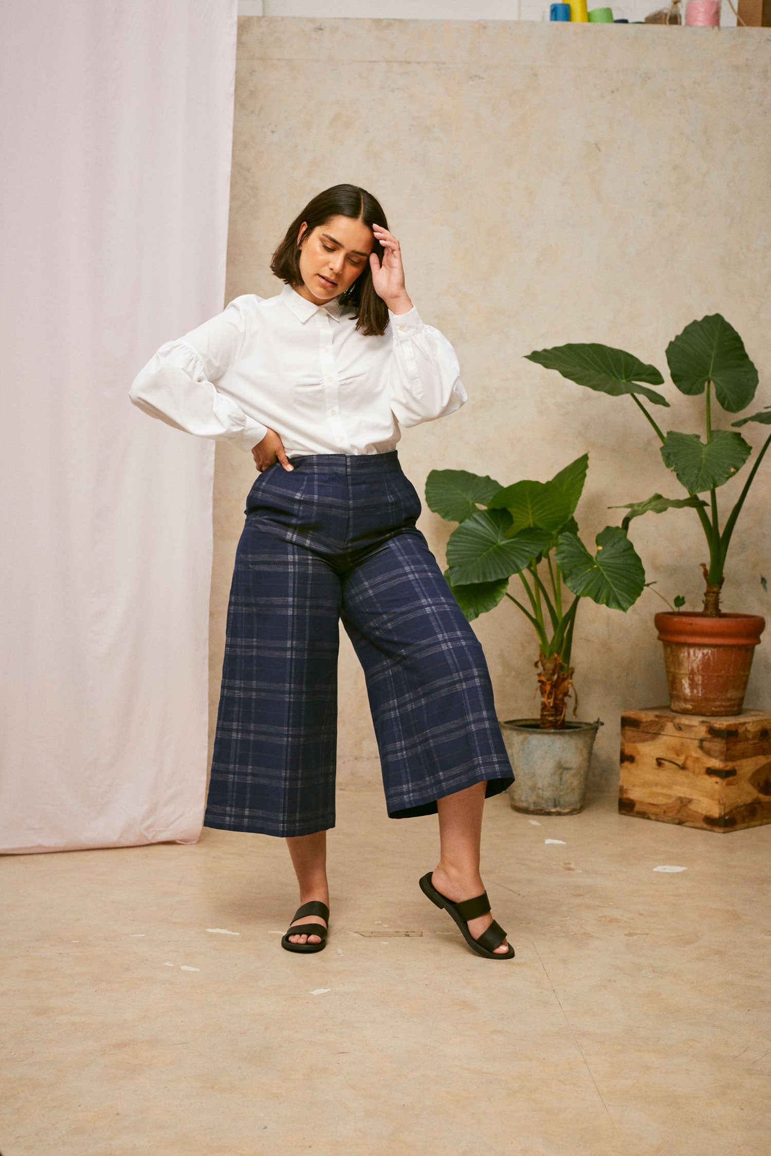 Full length shot of model wearing Saywood's Amelia check trousers, wide leg culotte shape in navy check, with black sandals. Worn with the white shirt, Edi volume sleeve shirt, tucked into the trouser. A plant and drop of pink fabric can be seen in the background.