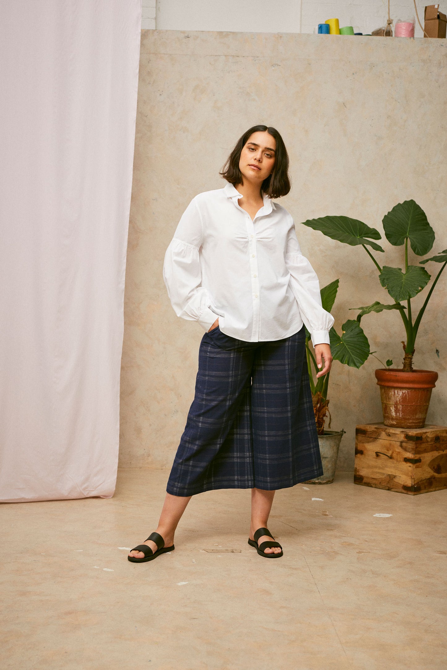 Full length shot of model wearing Saywood's Amelia check trousers, wide leg culotte shape in navy check, with black sandals. Worn with the white shirt, Edi volume sleeve shirt, tucked into the trouser. A plant and drop of pink fabric can be seen in the background.