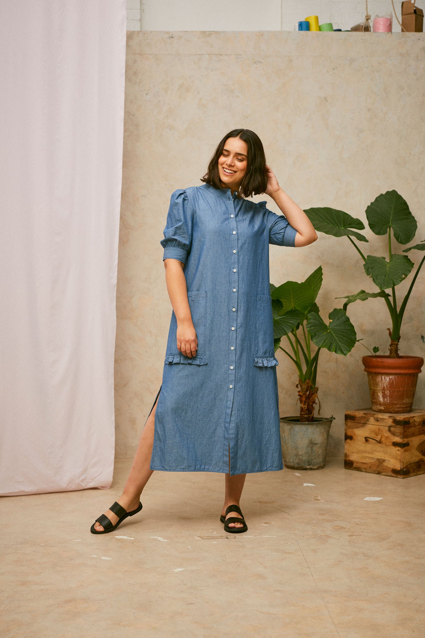 Full length shot of model wearing Saywood's Rosa denim shirtdress in light wash Japanese denim; a long line style with patch pockets and ruffles. Worn without belt and with black sandals. She smiles with her hand to the back of her head. A plant and drop of pink fabric can be seen in the background.