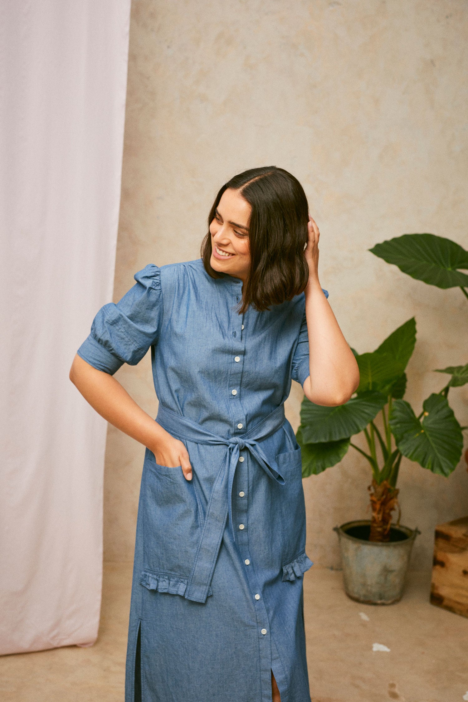 Model wears Saywood's Rosa denim shirtdress in light wash Japanese denim; a long line style with patch pockets and ruffles, a belt tied round the waist. She smiles with her hand to the back of her head. A plant and drop of pink fabric can be seen in the background.