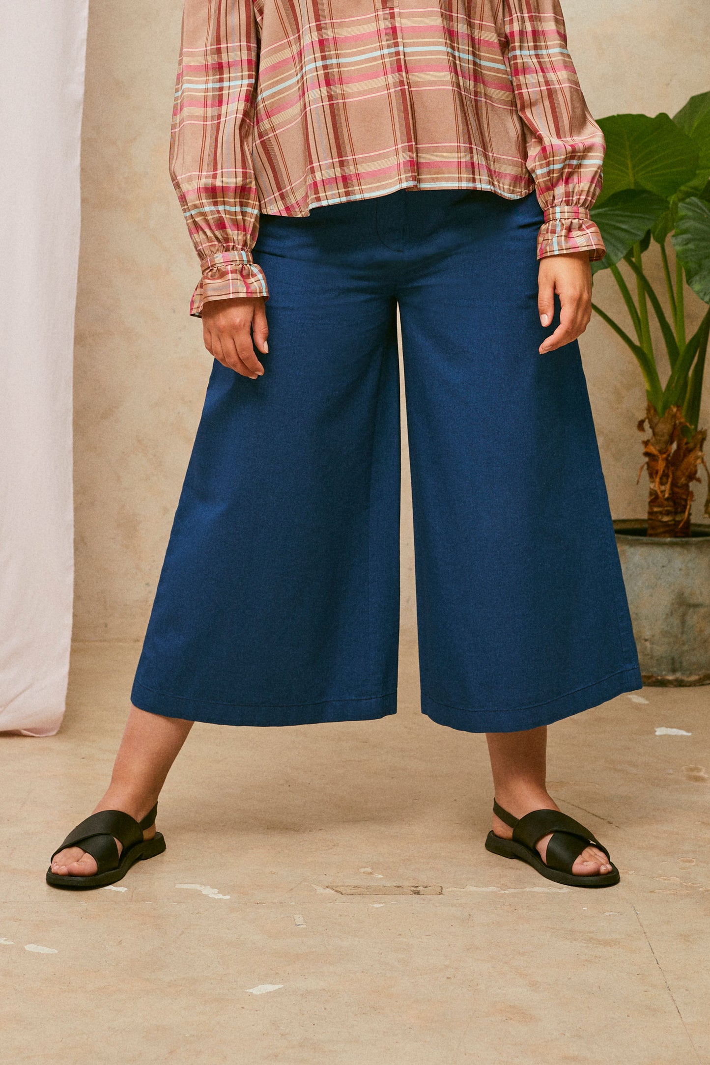 Close up of model wearing Saywood's Amelia denim trousers, wide leg culotte shape in Japanese denim, with black sandals. Worn with the pink check blouse, Marie a-line-shirt. A plant and drop of pink fabric can be seen in the background.
