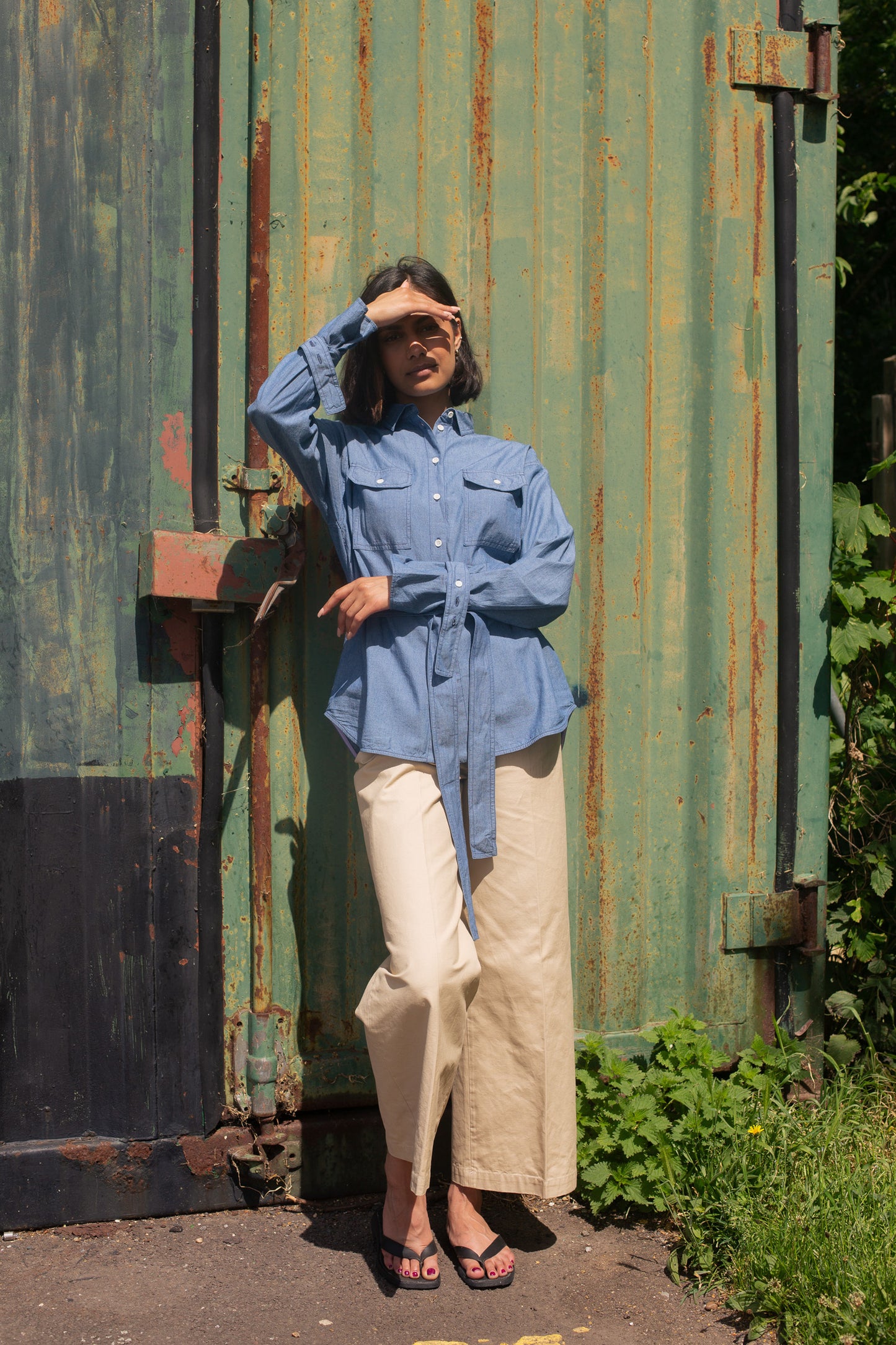 Model leans against a green shipping container, with one arm across her waist and the other to her forehead to block the sunlight. She wears Saywood's Zadie Boyfriend Shirt in Japanese Denim with a pair of beige trousers and black sandals.