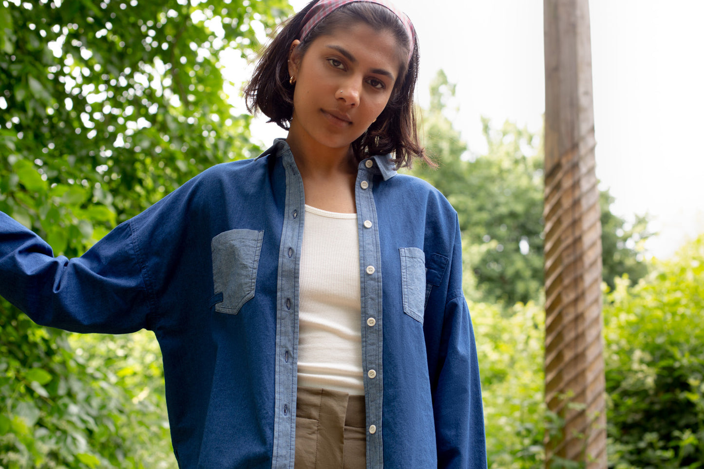 Model stands in surrounded by trees in the background. She wears Saywood's Japanese Denim Lela Patchwork Shirt, with light wash contrast cuffs, pockets and collar. Worn with khaki Amelia Trousers, with shirt left open with a white rib vest underneath.