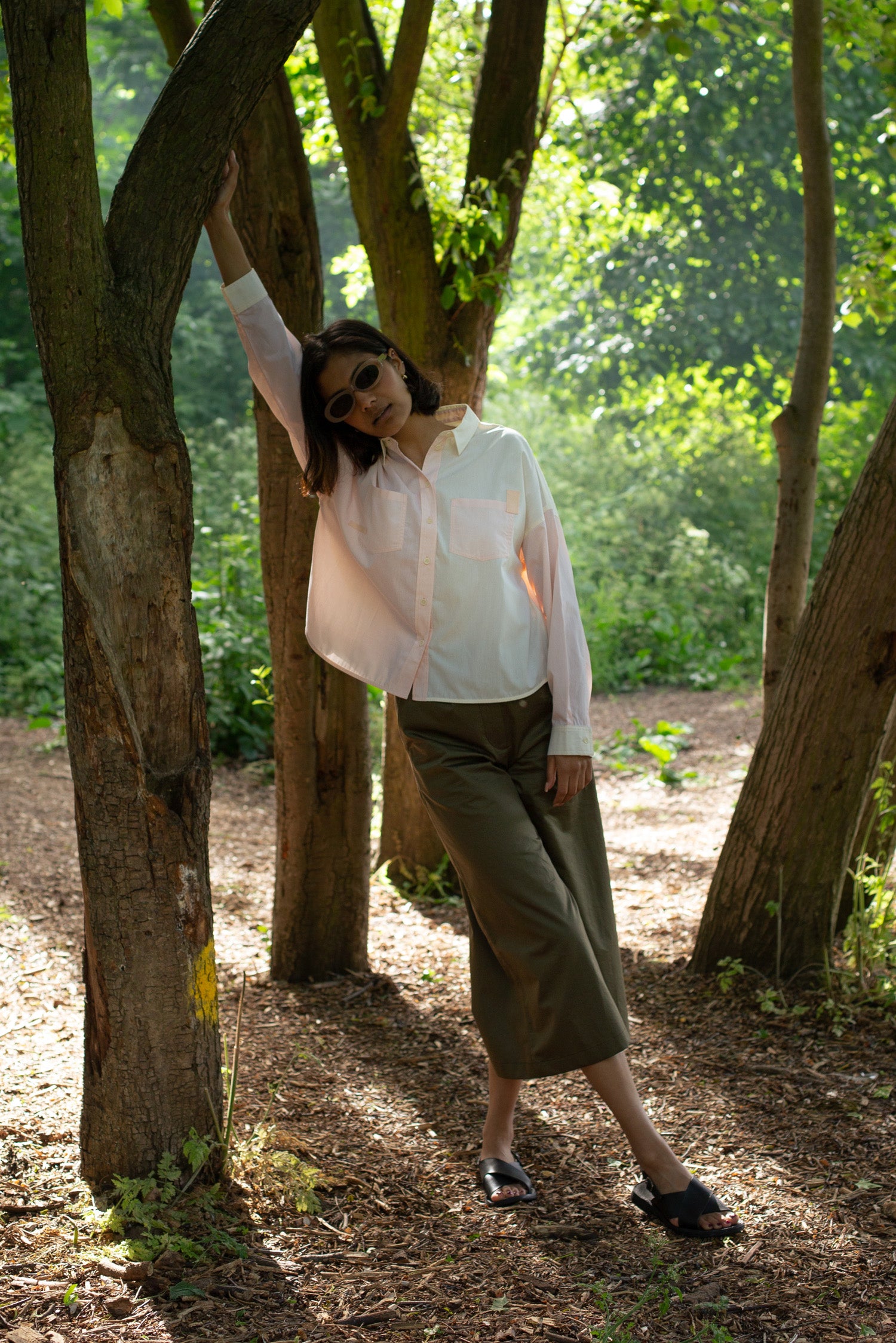 Model stands with one arm raised leaning against a tree, surrounded by nature. She wears the womens patchwork pastel shirt in orange and lemon, the Lela Shirt by Saywood. Boxy silhouette with patch pockets. Worn with the Khaki Amelia Trouser, sunglasses, and sandals.
