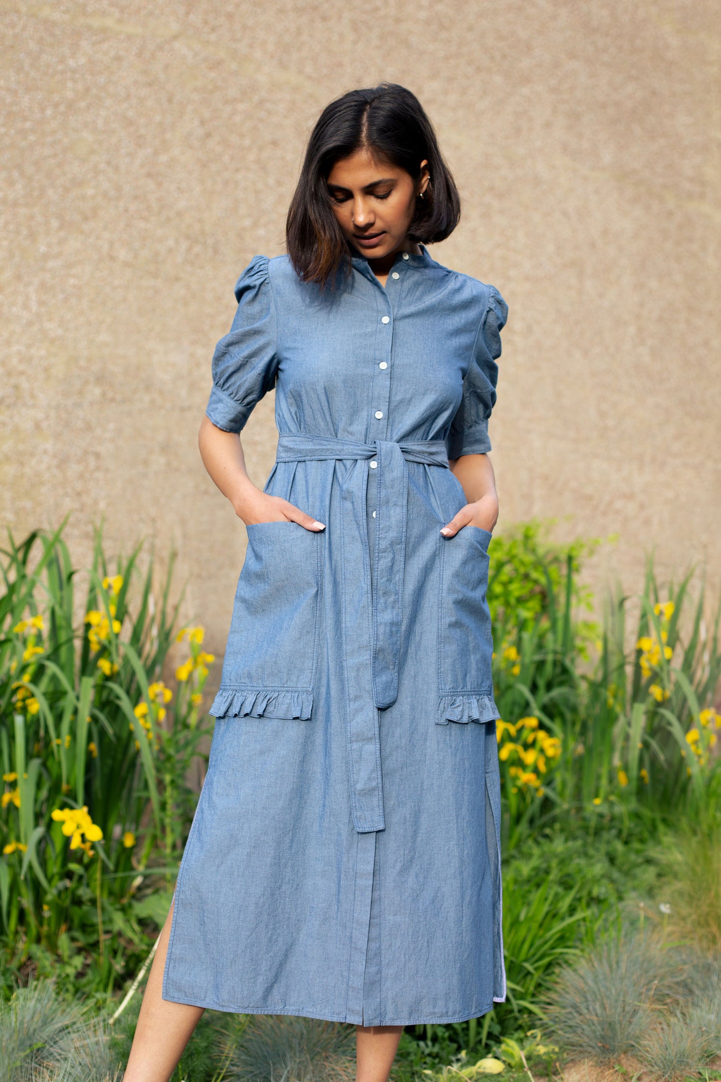 Model wears Saywood's Rosa denim shirtdress in light wash Japanese denim; a long line style with patch pockets and ruffles, a belt tied round the waist. She stands with her hands in her pockets loccking towards the floor. Yellow flowers can be seen in the background.