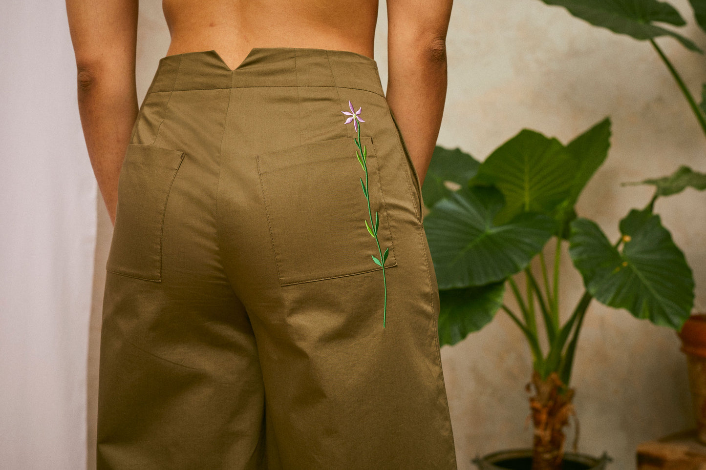 Cropped image of models wearing Saywood's Amelia khaki wide leg trouser; back waist view  showing patch pockets and Saywood signature flower embroidery in lilac. A plant and drop of pink fabric can be seen in the background.