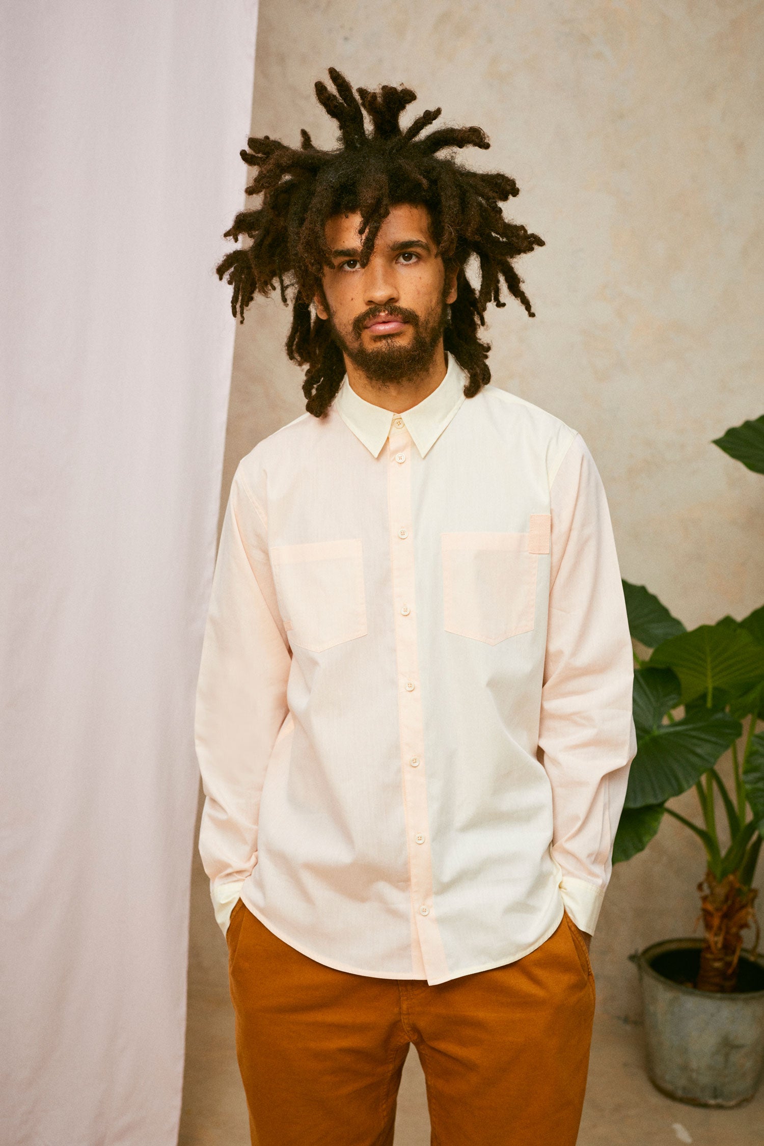 Model wears Saywood's Eddy Mens mens patchwork Shirt with patch pockets in orange and yellow. Worn with tabacco trousers. Model has both hands in his trouser pocket and a plant and drop of pink fabric can be seen in the background.