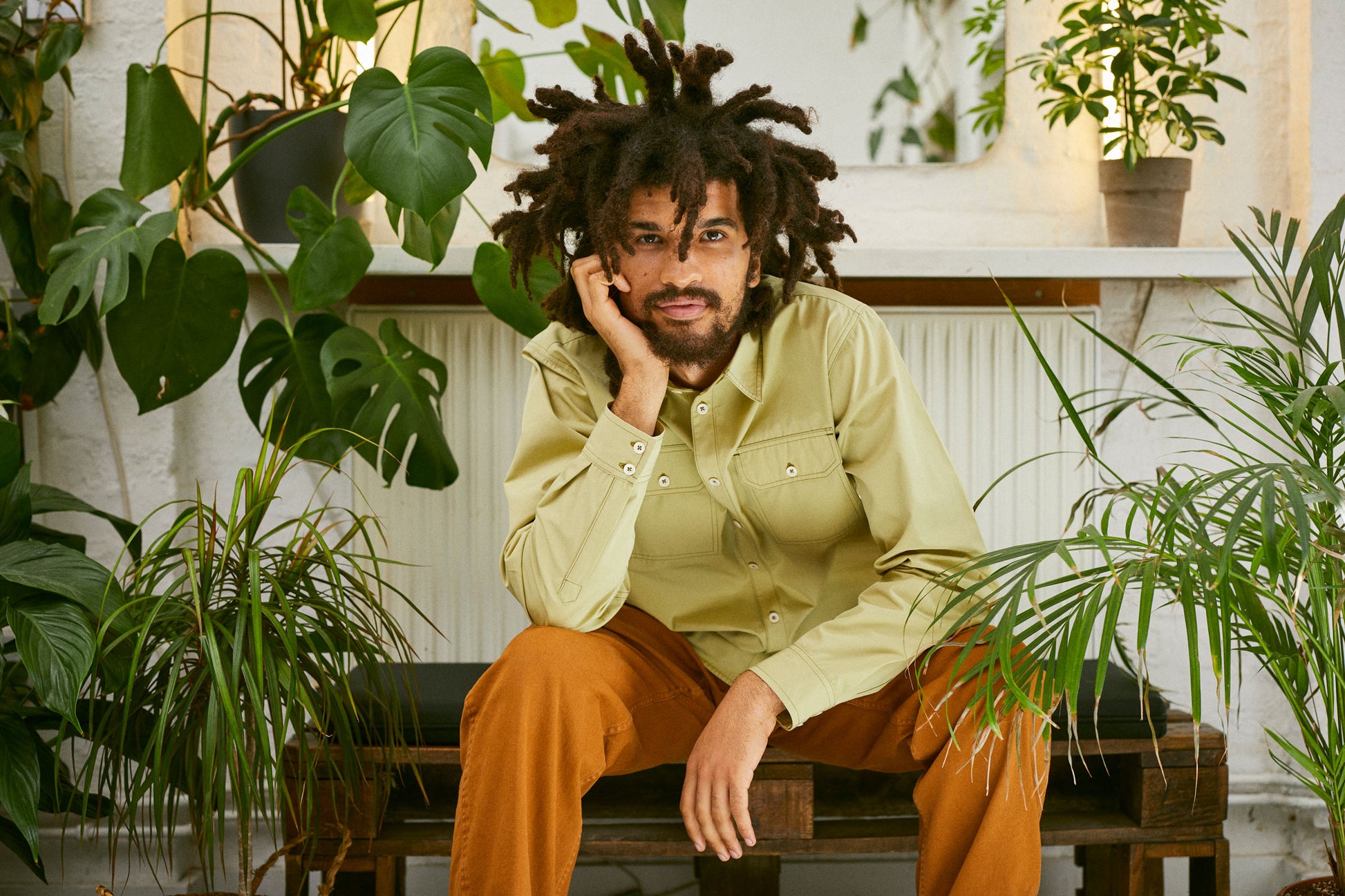 Model sits on a bench surrounded by house plants. One arm leans on his leg and his head leans on the other hand. He's wearing Saywood's Eddy Mens Khaki Shirt with utility pockets. Worn with tabacco trousers.