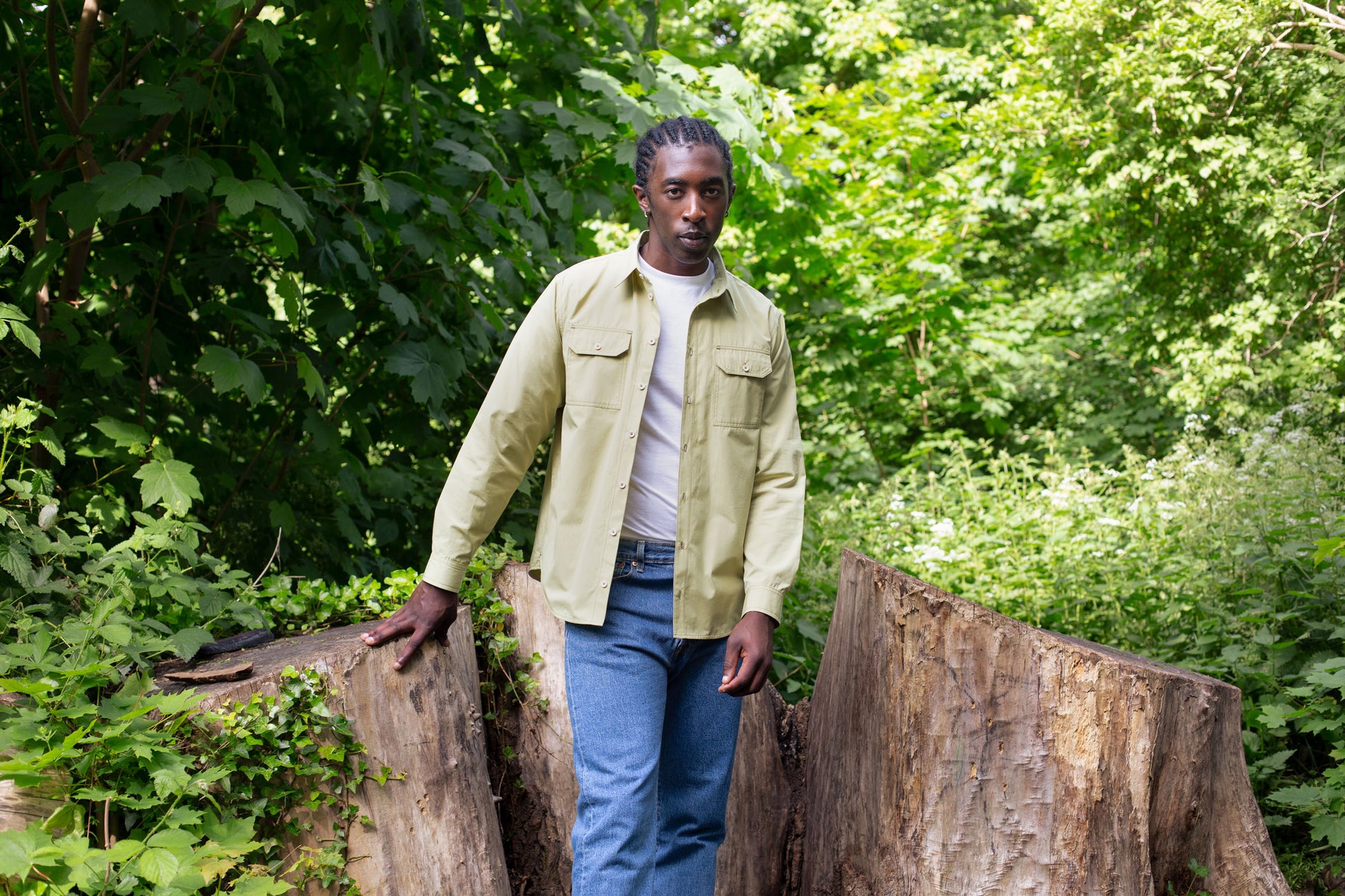 Model stands in front of several large tree stumps, with one hand rested on a stump, surrounded by greenery. He wears Saywood's Eddy Mens Khaki Shirt with utility pockets, worn open with a white t-shirt underneath. Worn with jeans.
