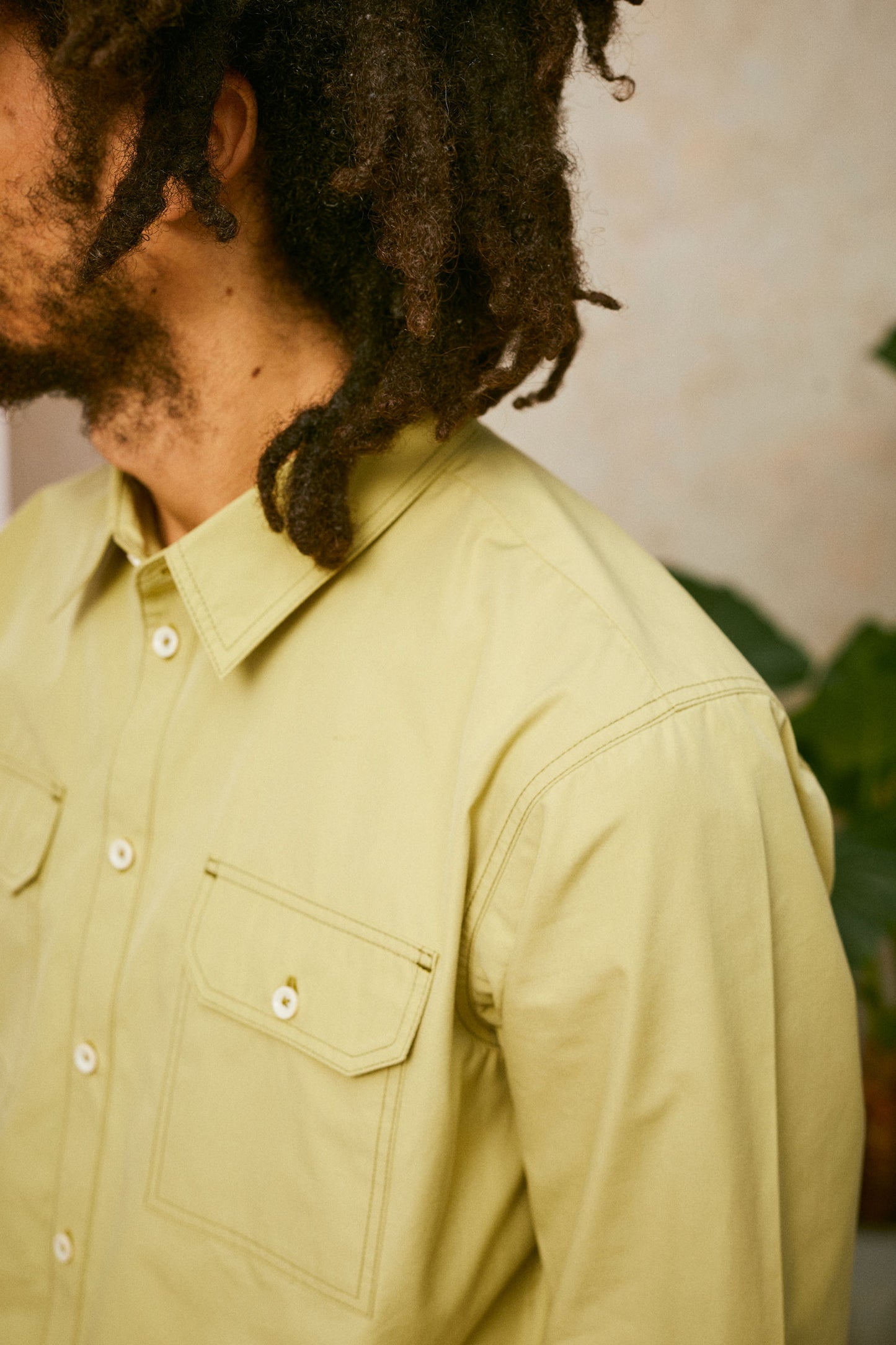 Close up of chest and shoulder; model wears Saywood's Eddy Mens Khaki Shirt. The images shows the utility pocket on the chest , collar and button placket. A plant can be seen in the background.