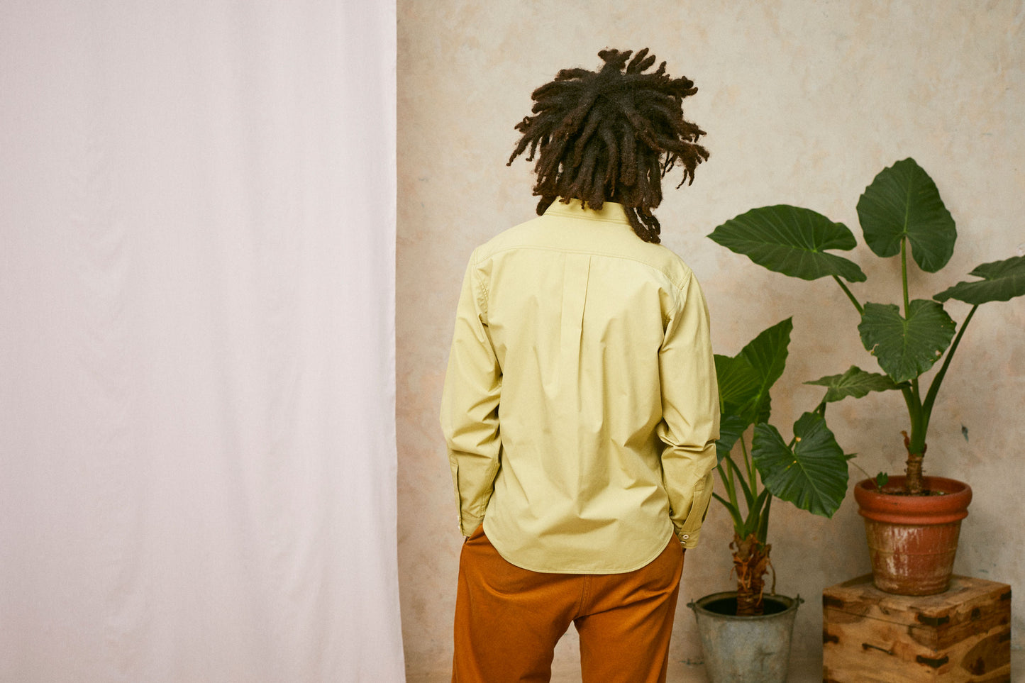 Close up back view of model wearing Saywood's Eddy Mens Khaki Shirt. Worn with tabacco trousers just visible. Model has both hands in his trouser pocket and a plant and drop of pink fabric can be seen in the background.
