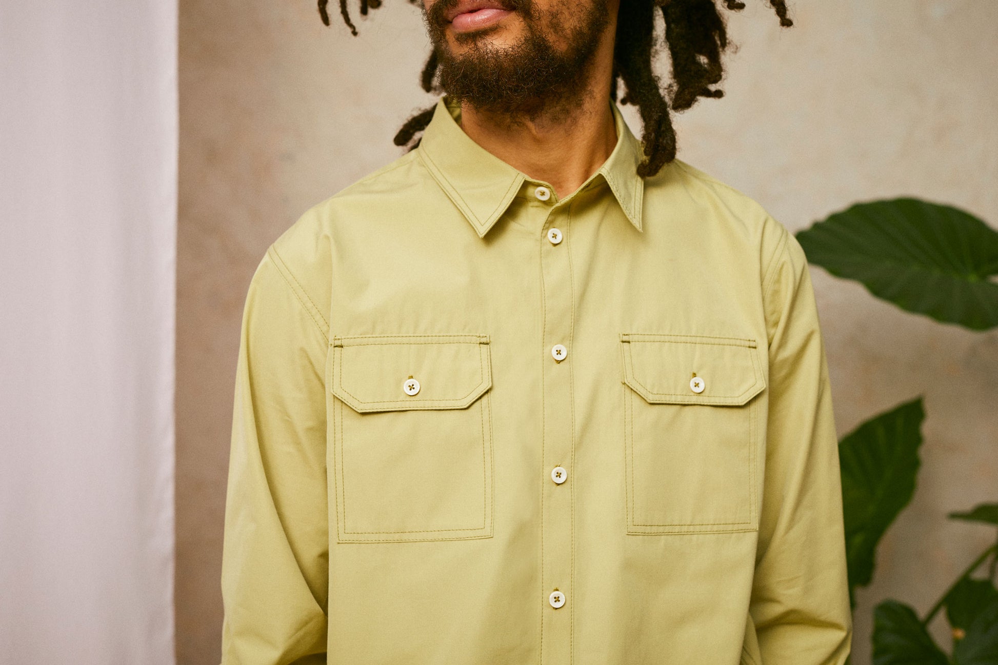 Close up of chest; model wears Saywood's Eddy Mens Khaki Shirt. The images shows the utility pockets on the chest and the top sticthing. A plant and drop of pink fabric can be seen in the background.