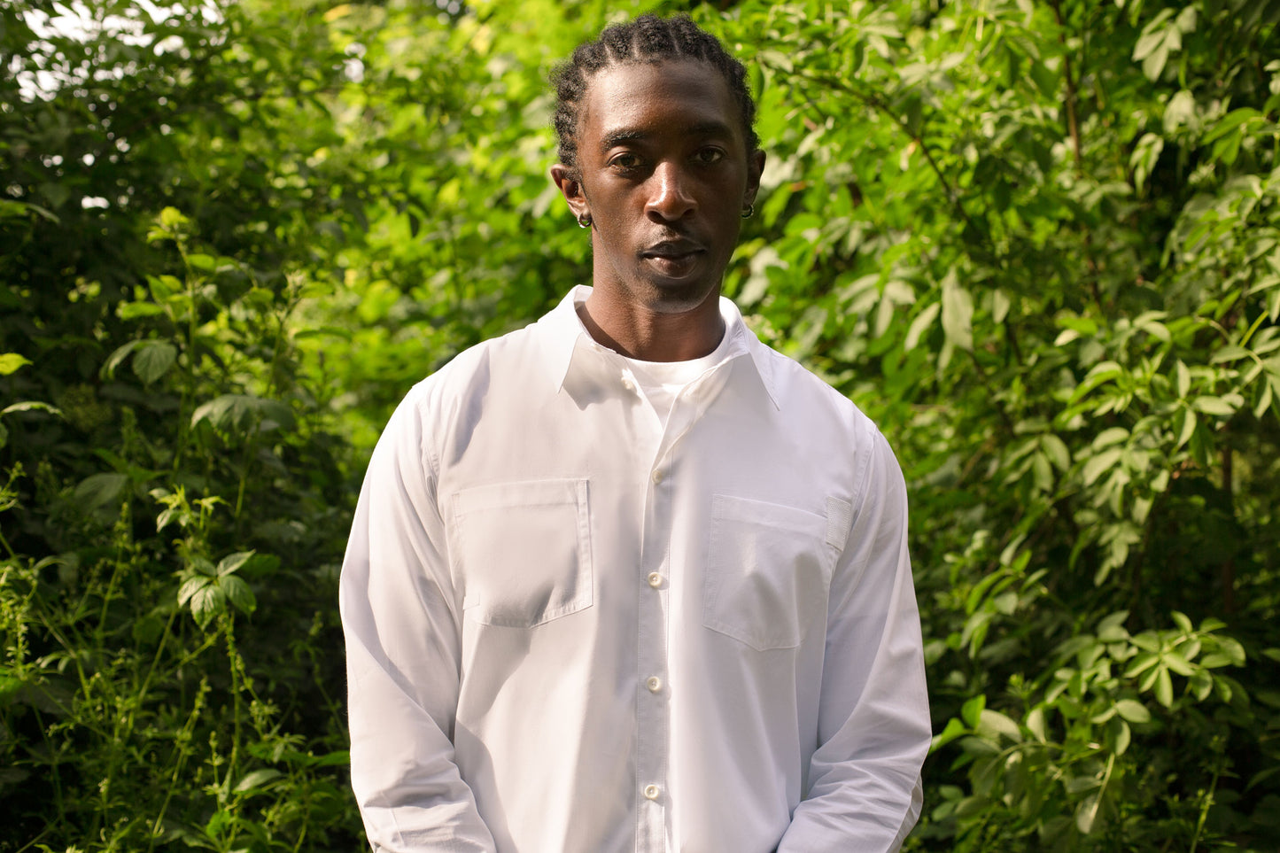 Close up of model wearing Saywood's Eddy Mens mens white Shirt with patch pockets, worn with the top buttons open and a white t-shirt underneath. The model is surrounded by trees and greenery.