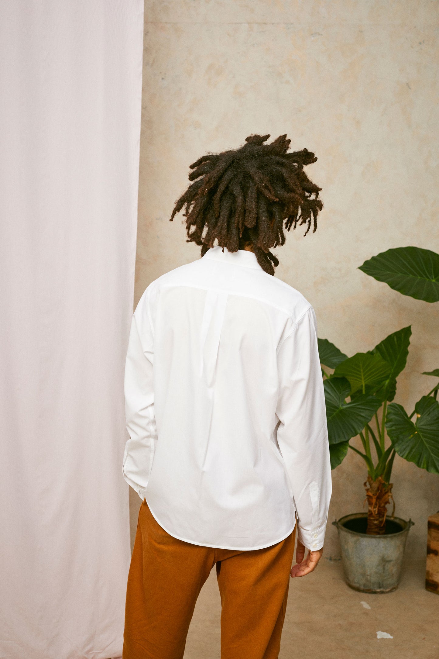 Back view of model wearing Saywood's Eddy Mens mens white Shirt with patch pockets; showing the back yoke and box pleat in the back. Worn with tabacco trousers. Model has one hand in his trouser pocket. A plant and drop of pink fabric can be seen in the background.
