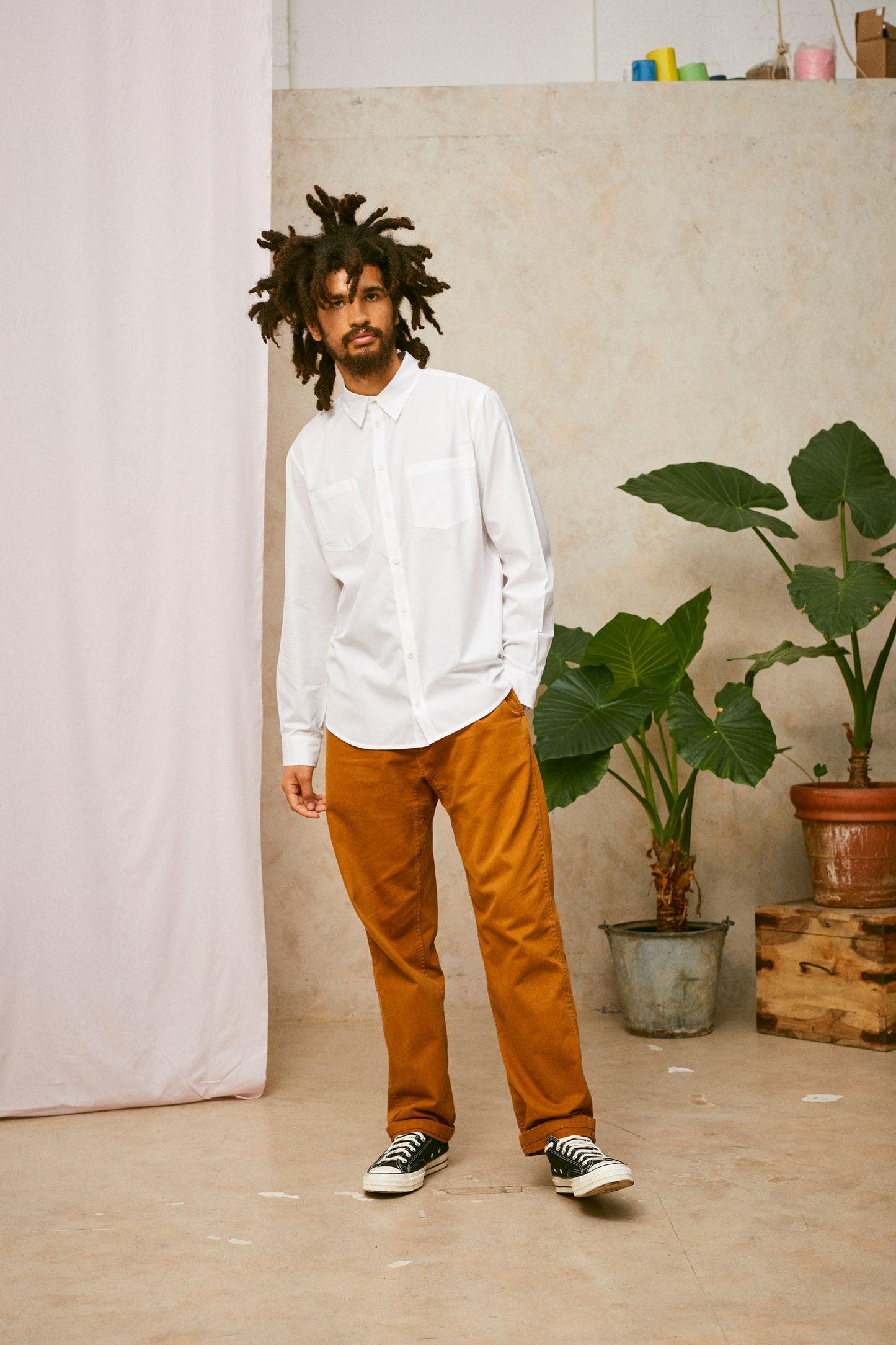 Full length shot of model wearing Saywood's Eddy Mens mens white Shirt with patch pockets. Worn with tabacco trousers and black Converse. Model has one hand in his trouser pocket. A plant and drop of pink fabric can be seen in the background.