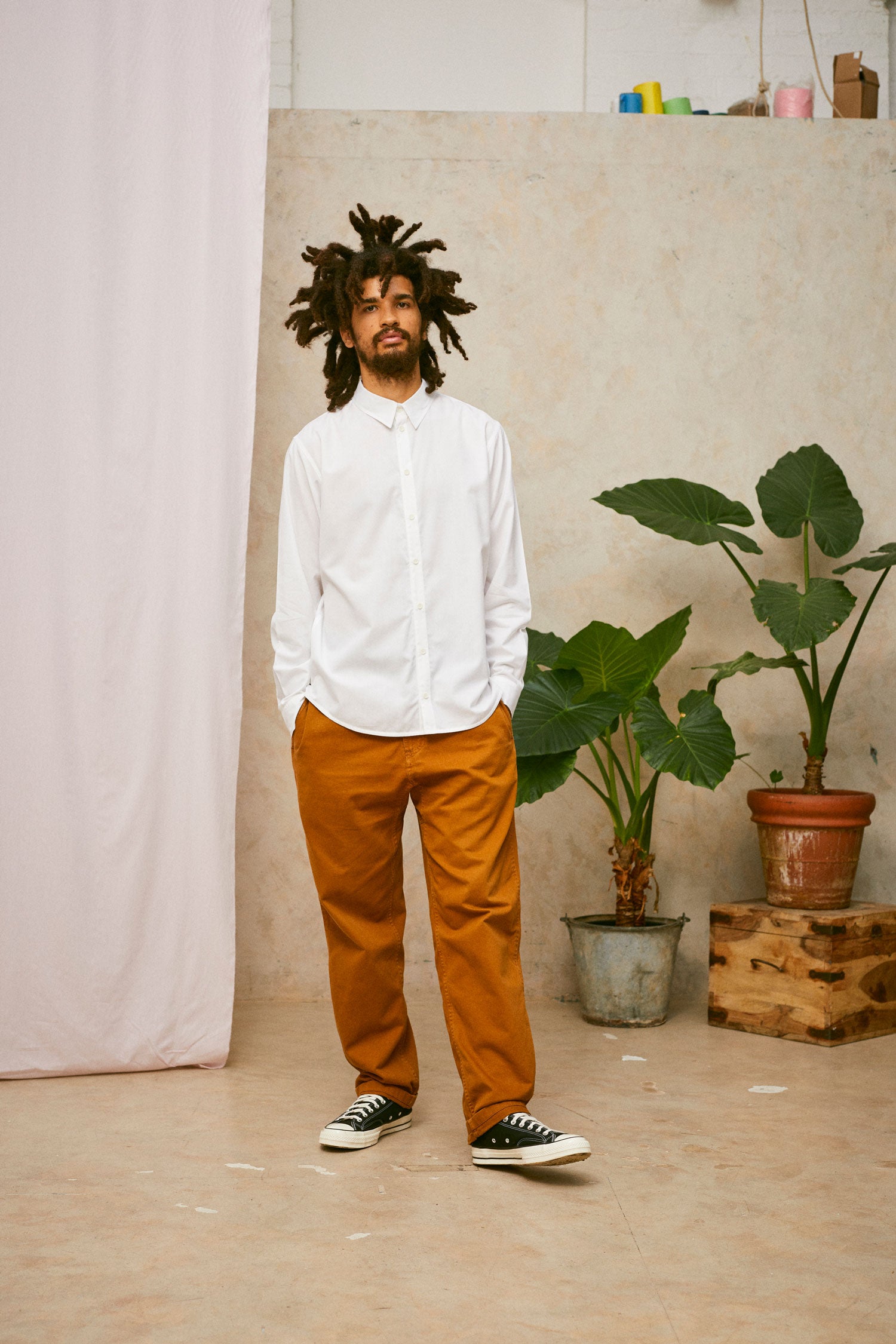 Full length shot of model wearing Saywood's Eddy Mens mens white Shirt. Worn with tabacco trousers and black Converse. Model has both hands in his trouser pocket and a plant and drop of pink fabric can be seen in the background.