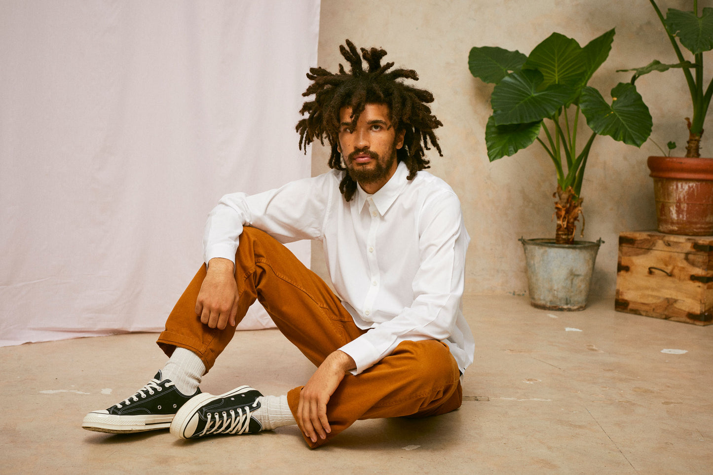 Model sits on the floor, with one knee up and his arm resting on his knee. He is wearing Saywood's Eddy Mens mens white Shirt. Worn with tabacco trousers and black Converse. A plant and drop of pink fabric can be seen in the background.