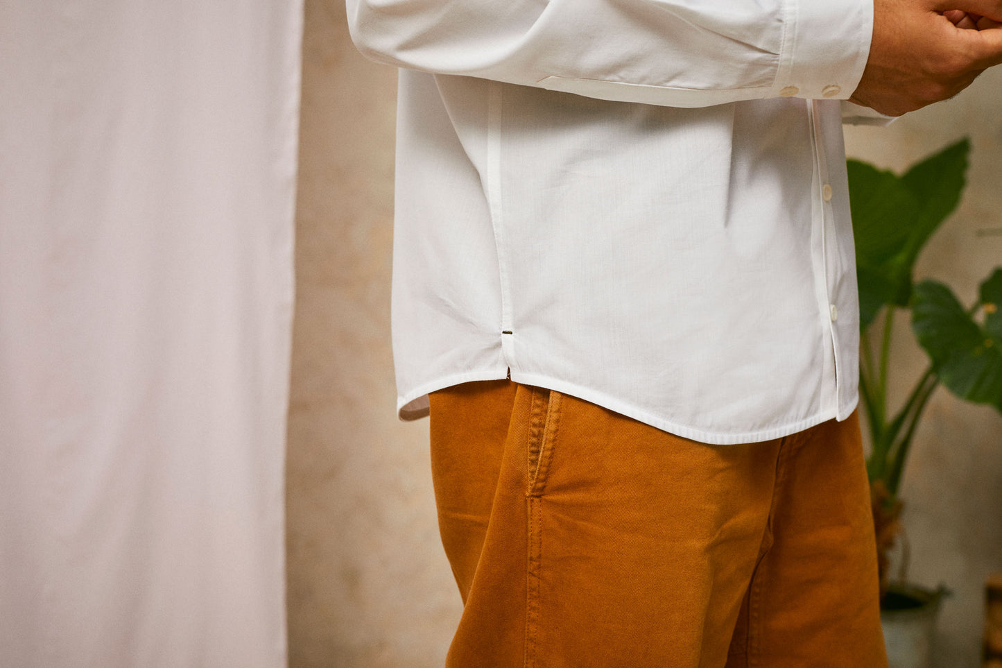 Close up of model's hips, wearing Saywood's Eddy Mens mens white Shirt. Worn with tabacco trousers just visible. Model holds both hands in front of his waist, showing the side split and contrast dark green bar tack at the side hem. A plant and drop of pink fabric can be seen in the background.