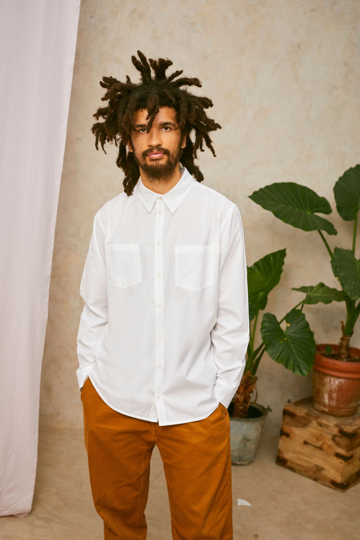 Model wears Saywood's Eddy Mens mens white Shirt with patch pockets. Worn with tabacco trousers. Model has both hands in his trouser pocket and a plant and drop of pink fabric can be seen in the background.