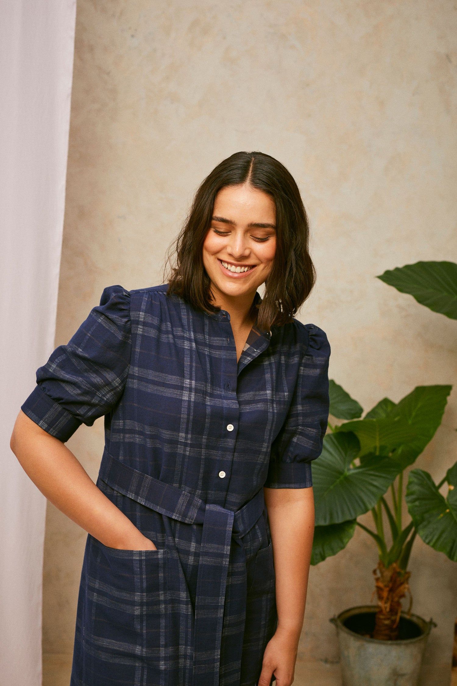 Close up crop of model wearing Saywood's Rosa navy check shirtdress, with belt loosely tied round the waist. The model is smiling and a plant and drop of pink fabric can be seen in the background.