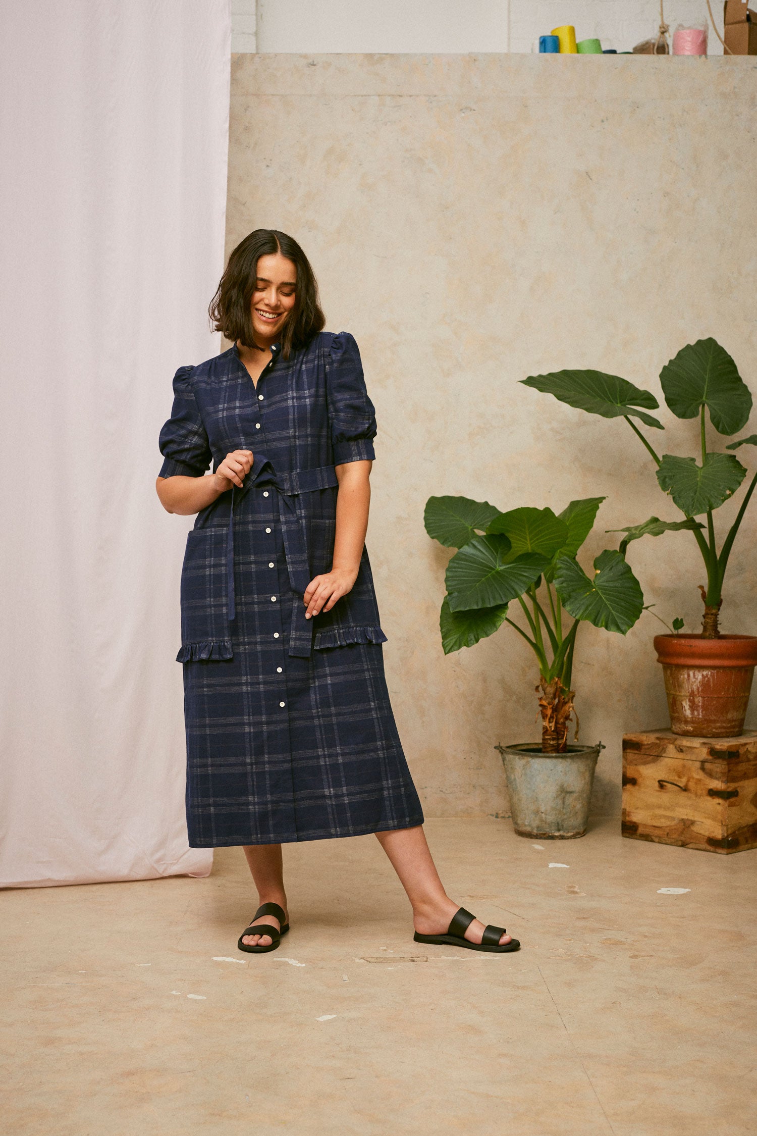 Full length shot of model wearing Saywood's Rosa navy check shirtdress, with belt tied round the waist. The model is smiling and pulling at the belt ties. Worn with black sandals. A plant and drop of pink fabric can be seen in the background.