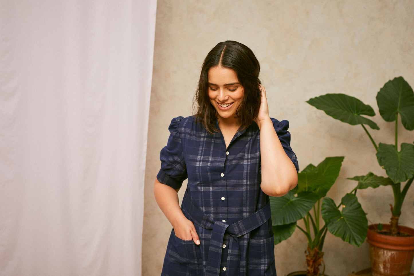 Close up crop of model wearing Saywood's Rosa navy check shirtdress, with belt loosely tied round the waist. The model is smiling with one hand to the back of her head, and a plant and drop of pink fabric can be seen in the background.