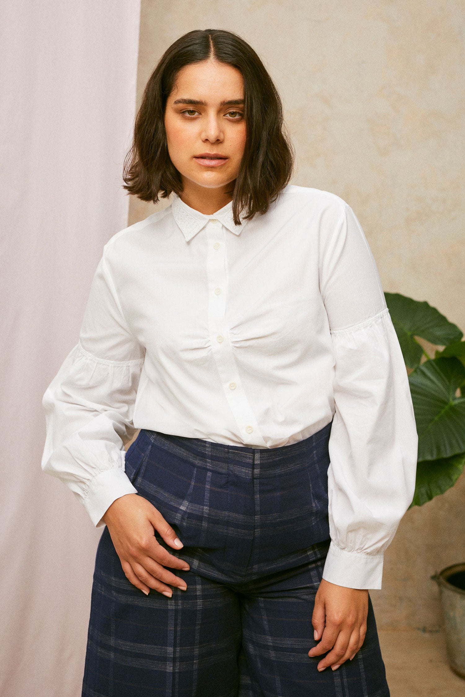 Close up of model wearing Saywood's Amelia check trousers, wide leg culotte shape in navy check. Worn with the white shirt, Edi volume sleeve shirt with lace collar, tucked into the trouser. A plant and drop of pink fabric can be seen in the background.