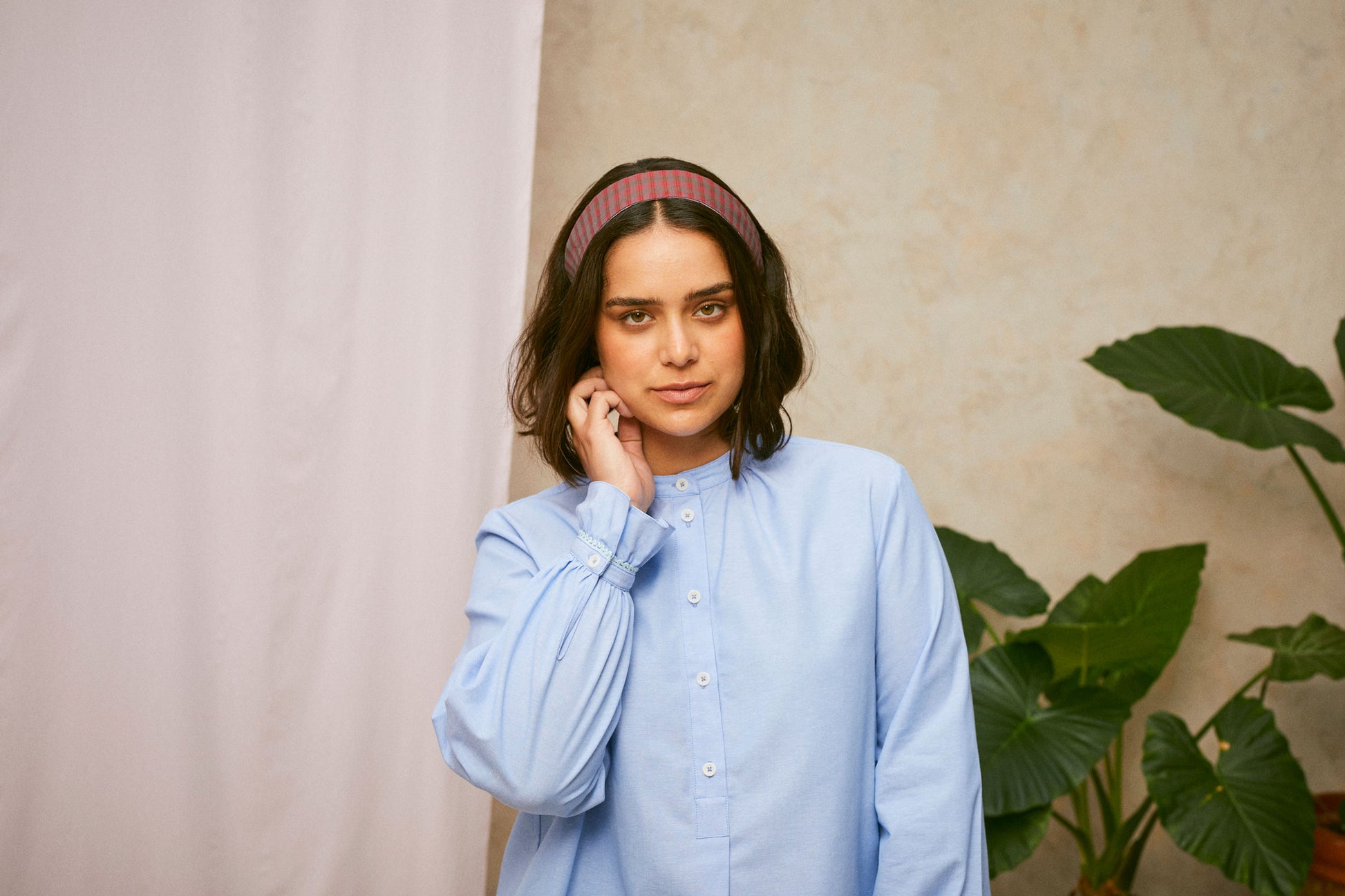 Close up of model wearing Saywood's pale blue shirt, the Marie A-Line Blouse, with the red check Heidi Headband worn in her her. The lace trim frill cuff can be seen as she holds her hand to her face. A plant and drop of pink fabric can be seen in the background. 