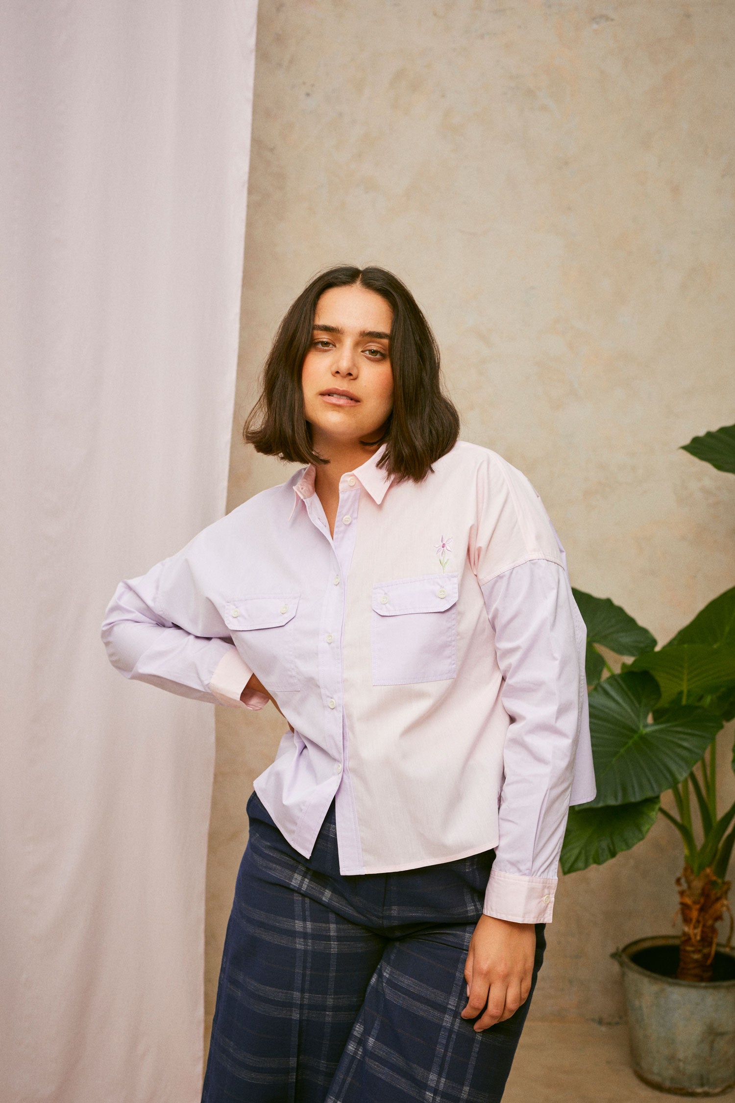 Model wears the Jules Utility Shirt: colourblocked pink and lilac shirt with a boxy silhouette. Jules Shirt from Saywood with embroidered flower detail and utility style pockets. Worn with a navy check trouser, she stands in front of a pink fabric drop and a plant.